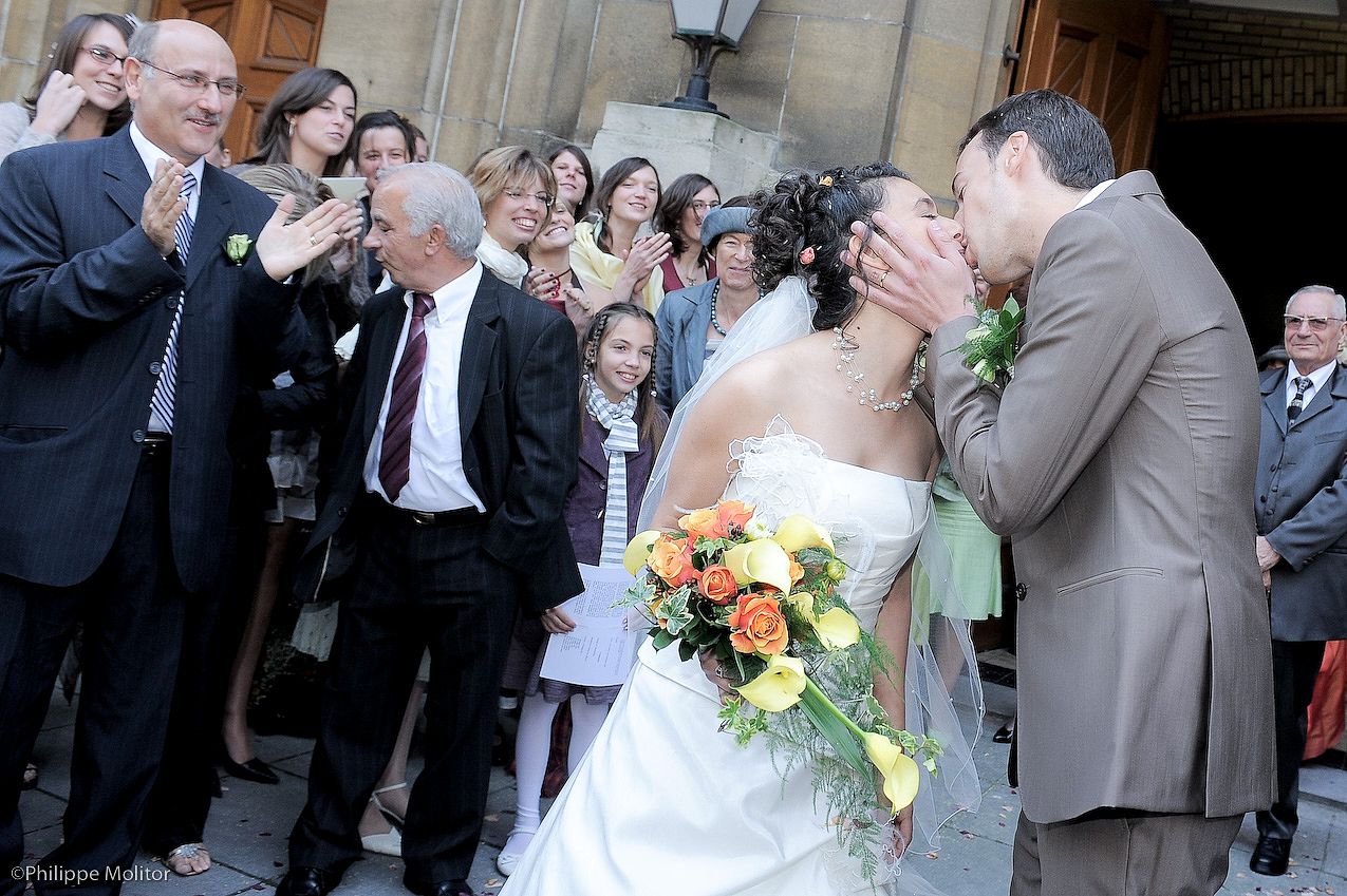 Bride and groom share a kiss outside church surrounded by cheering wedding guests, bride holding colorful bouquet.