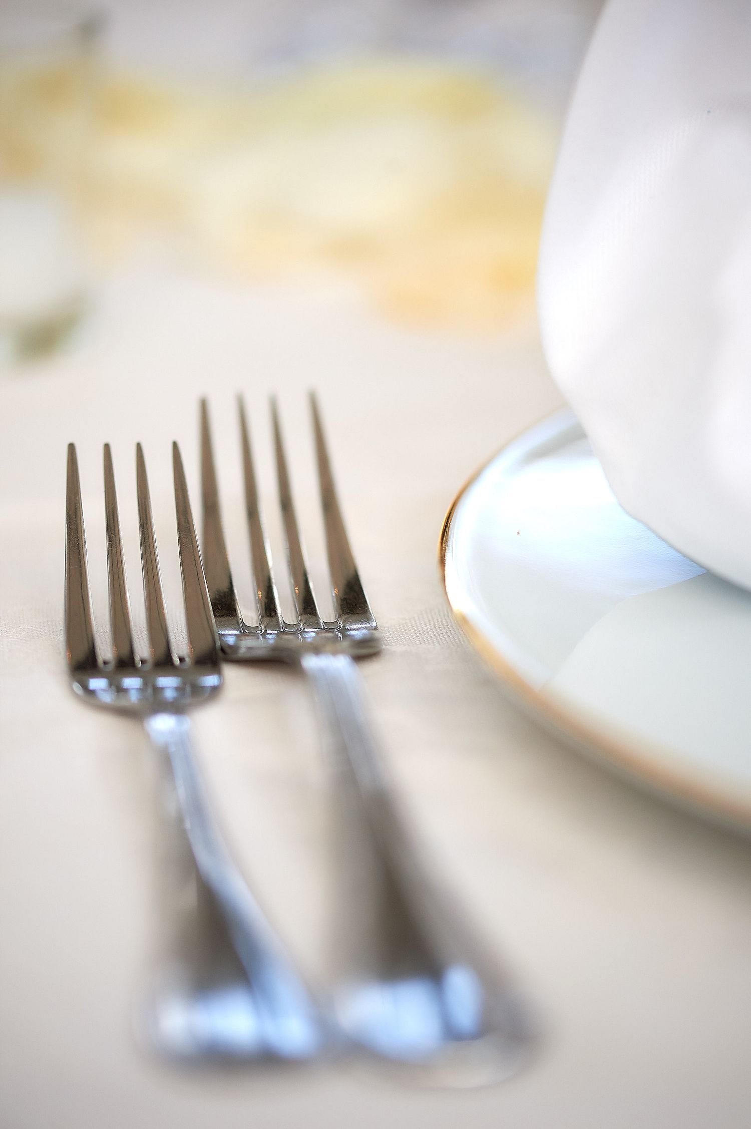 Two silver forks beside a white china cup and saucer with gold trim on an elegant table setting.