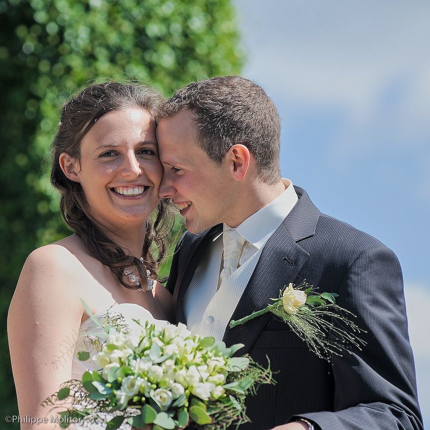 Happy bride and groom embracing outdoors, bride holding white flower bouquet, groom wearing suit with boutonniere on sunny day.