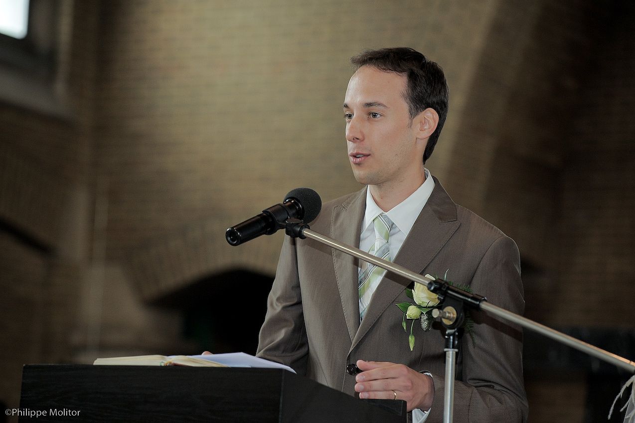 Young man in brown suit speaking at podium with microphone during formal ceremony or wedding reception.