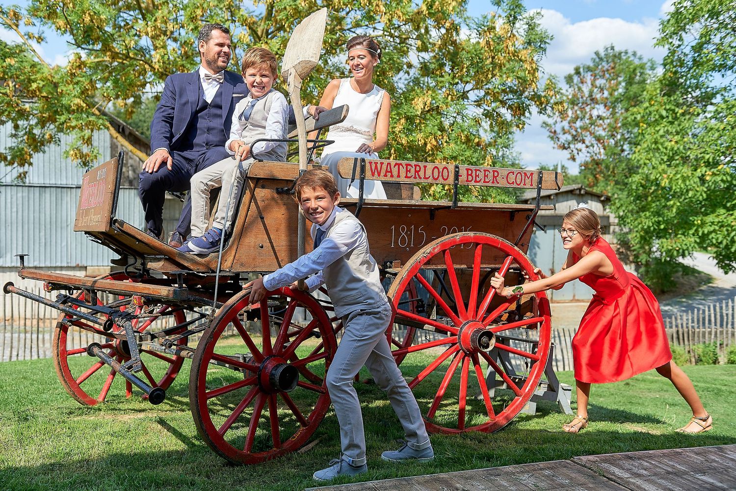 Family posing playfully on a vintage red Waterloo horse-drawn wagon outdoors on a sunny day.