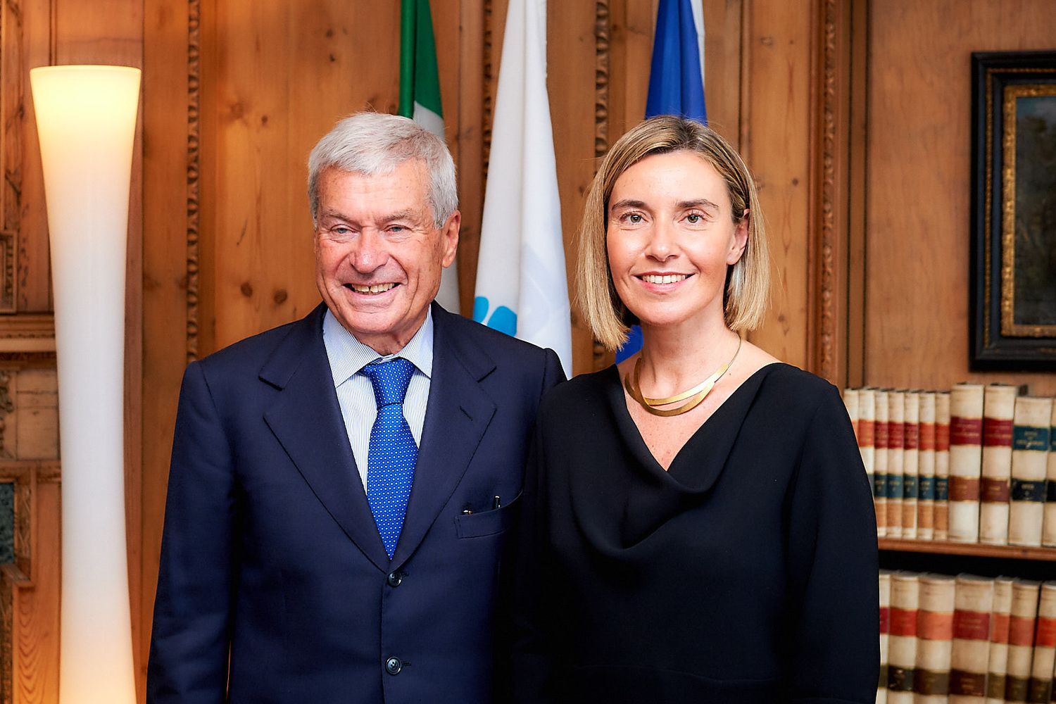 Two people in formal business attire pose together in front of flags in an official setting.