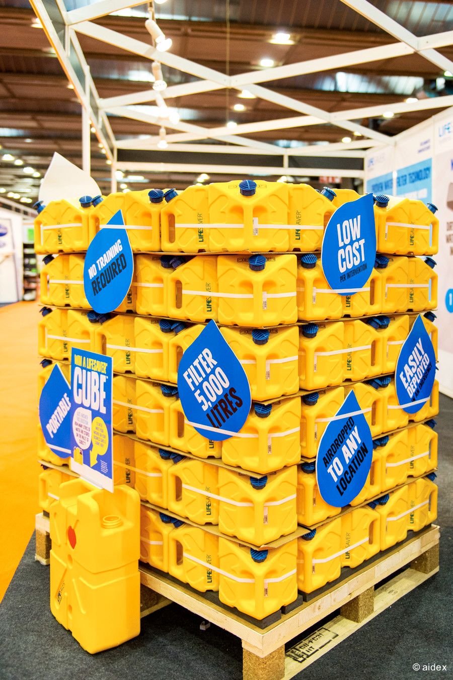 Stack of yellow plastic containers with blue promotional tags on a wooden pallet in a retail setting.