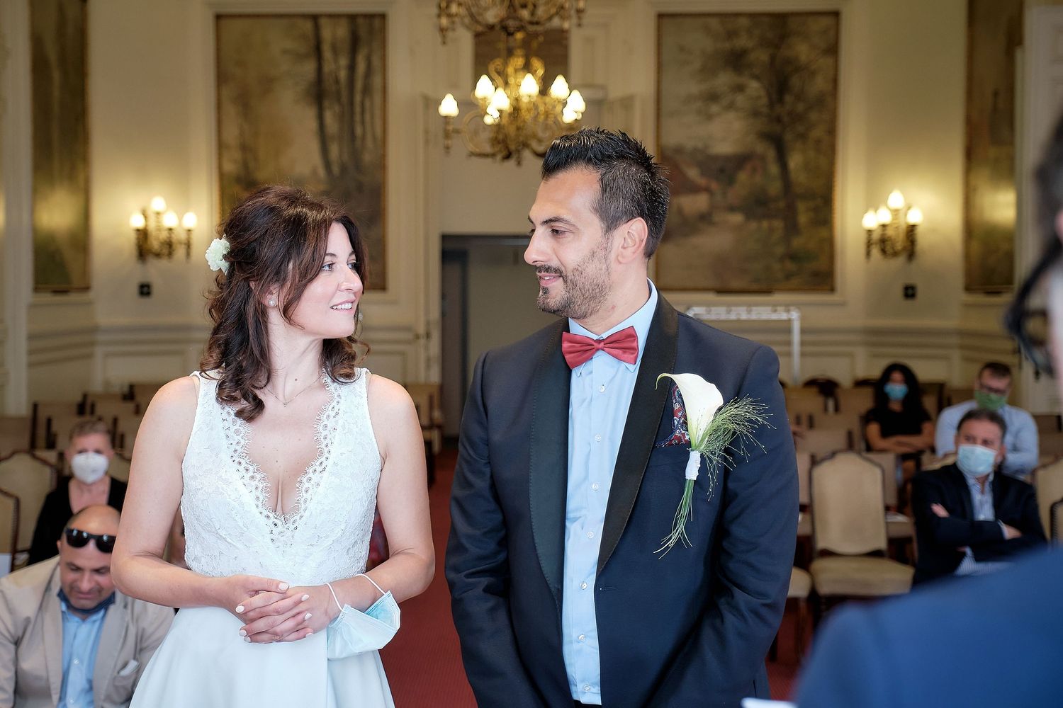 Bride and groom exchange smiles during elegant wedding ceremony in ornate hall with guests wearing masks.
