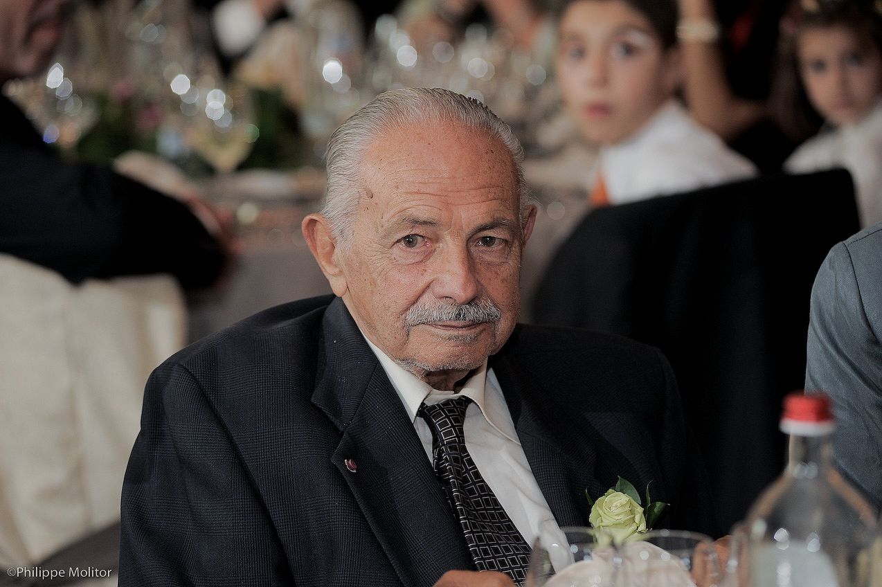 Elderly man with mustache in dark suit and tie seated at formal event dinner table.
