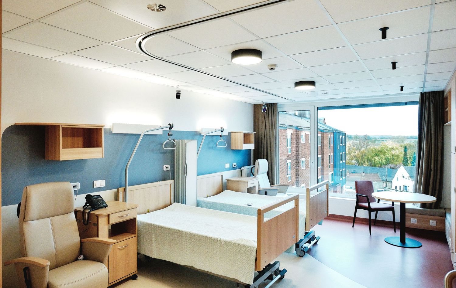 Wide angle view of hospital room showing bed, seating area and large windows.