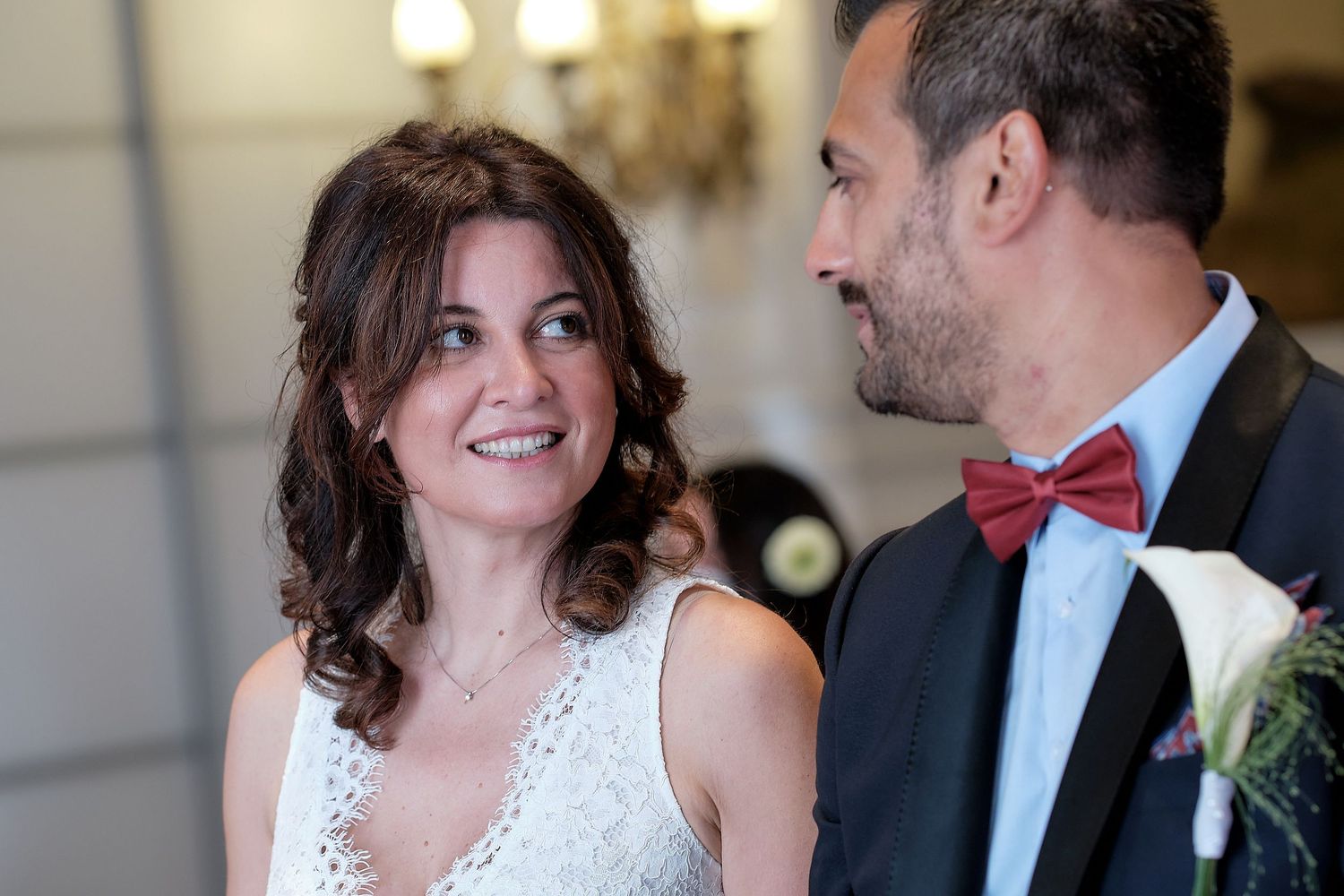 A bride in a white dress gazes lovingly at a groom wearing a blue suit and red bow tie at a wedding ceremony.
