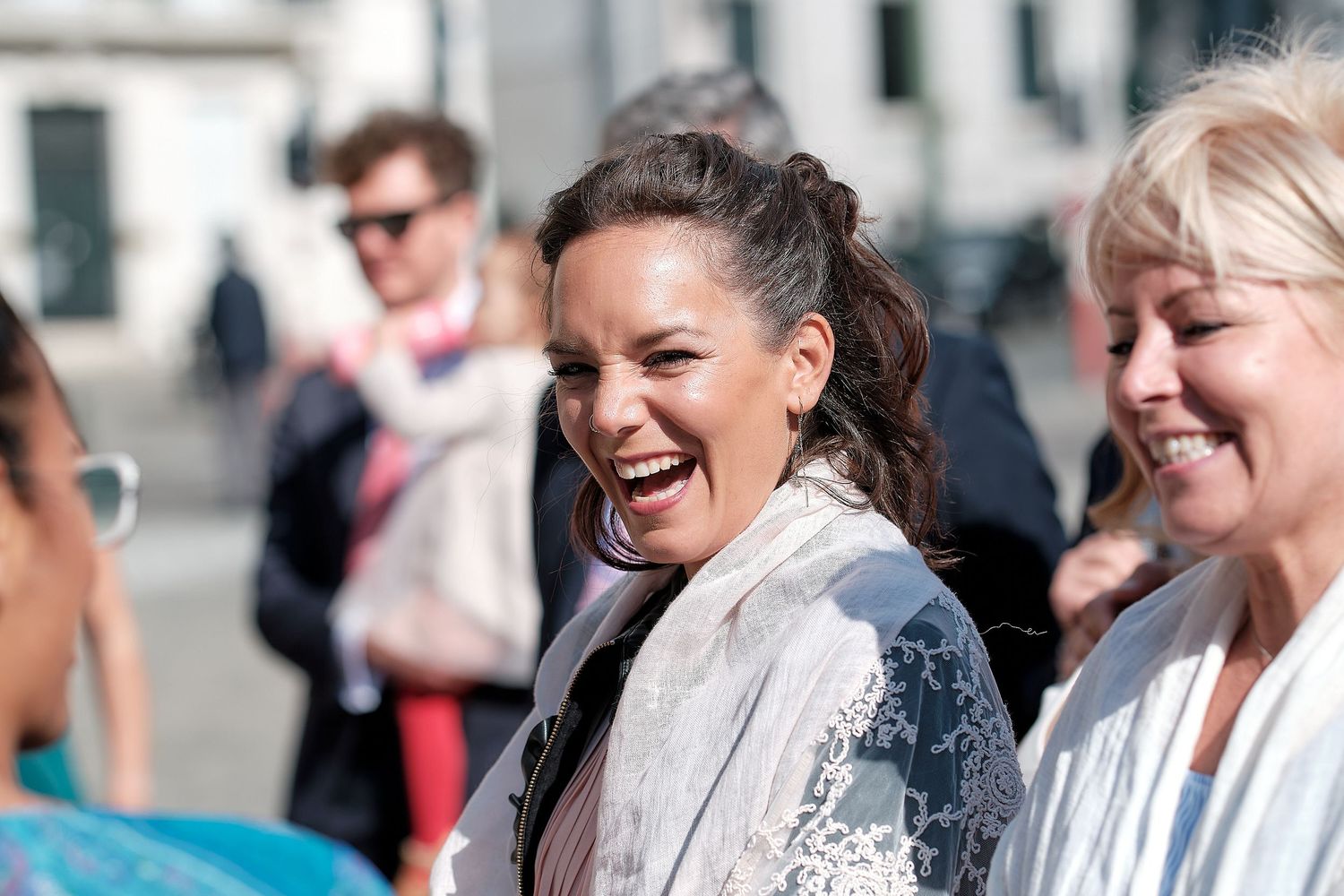A woman laughs joyfully outdoors with smiling people in the background on a sunny day.