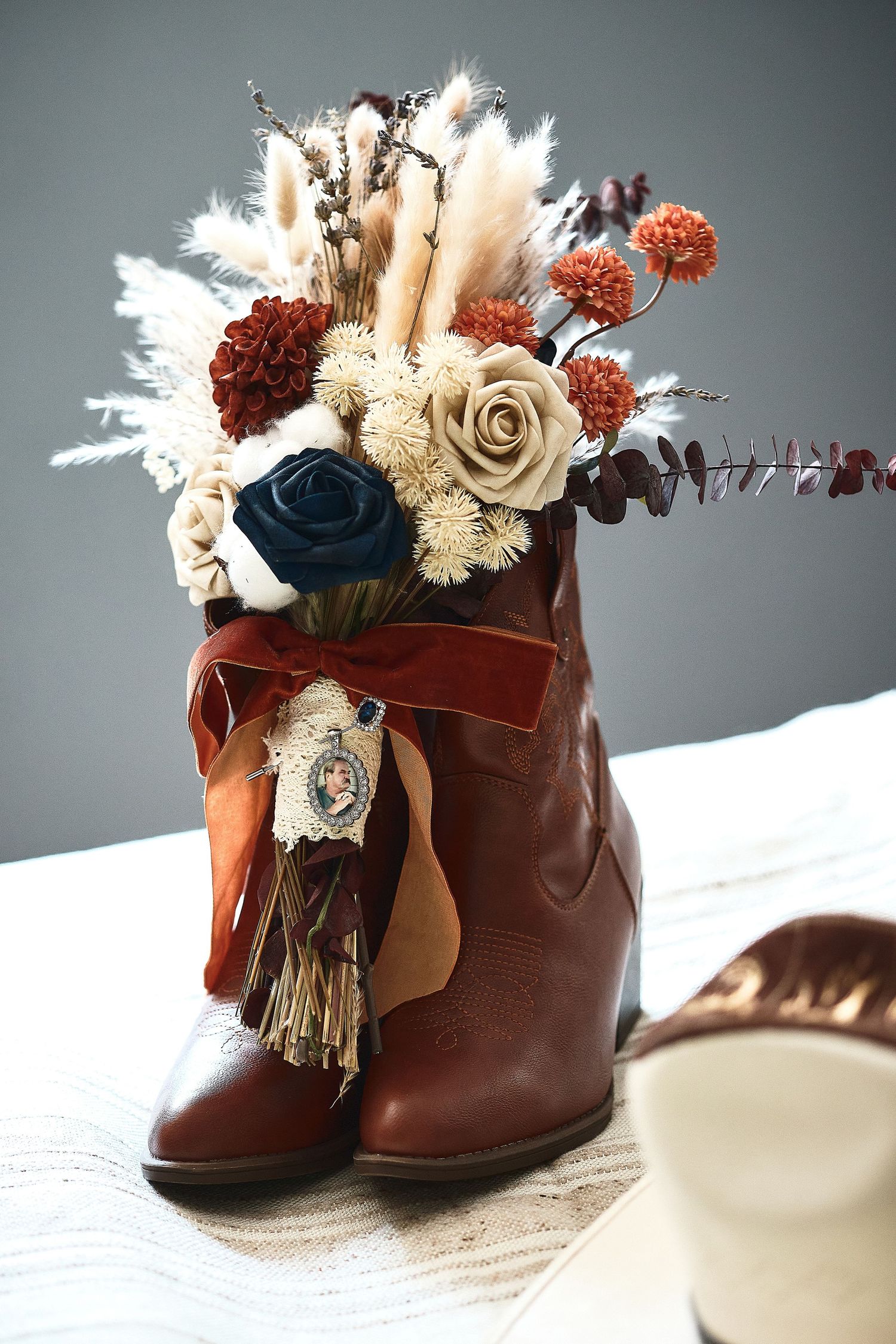 Brown cowboy boot vase with dried floral arrangement featuring roses, pampas grass, and burgundy ribbon on white surface.