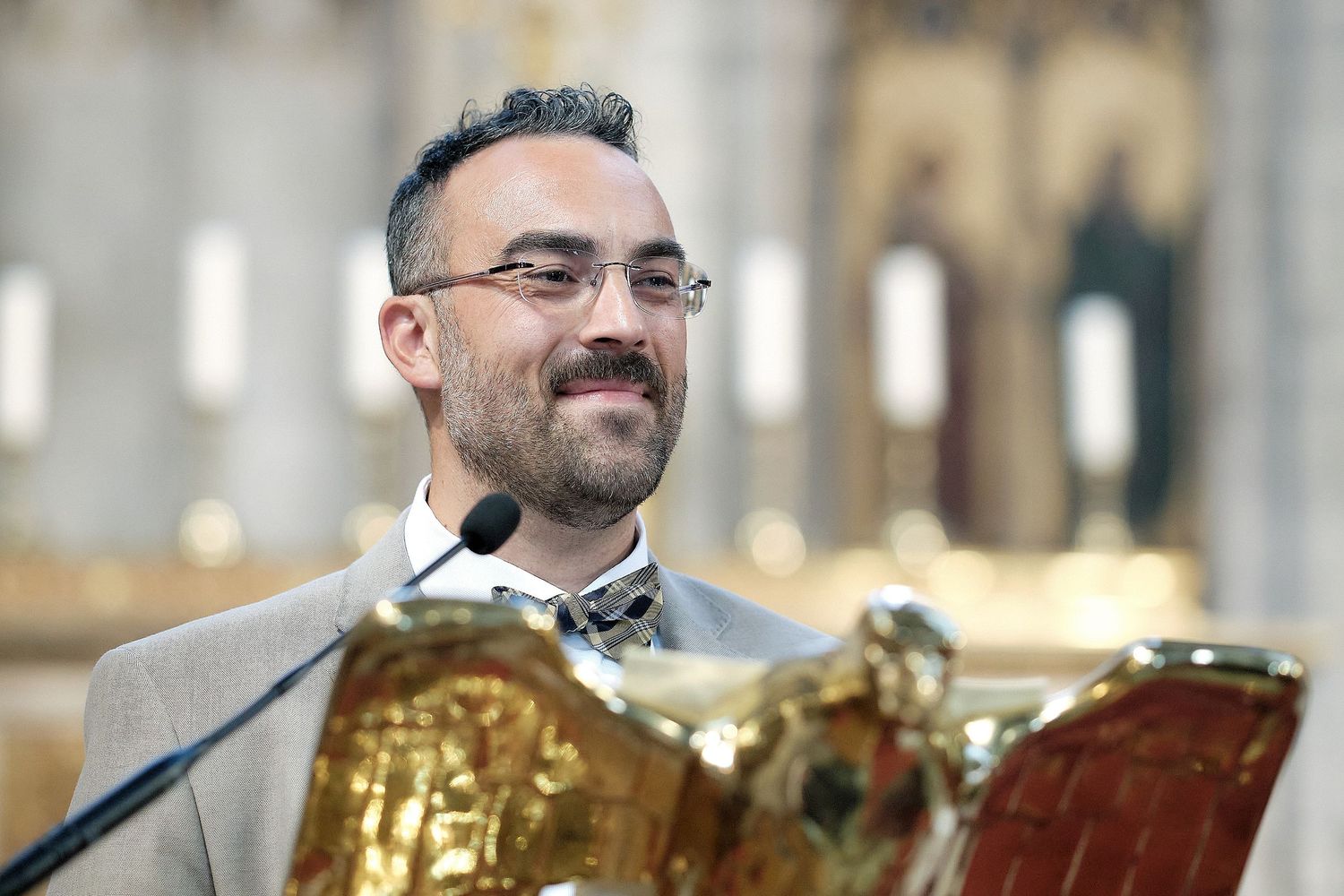 Smiling bearded man in suit with bow tie speaking at ornate golden podium in grand cathedral.