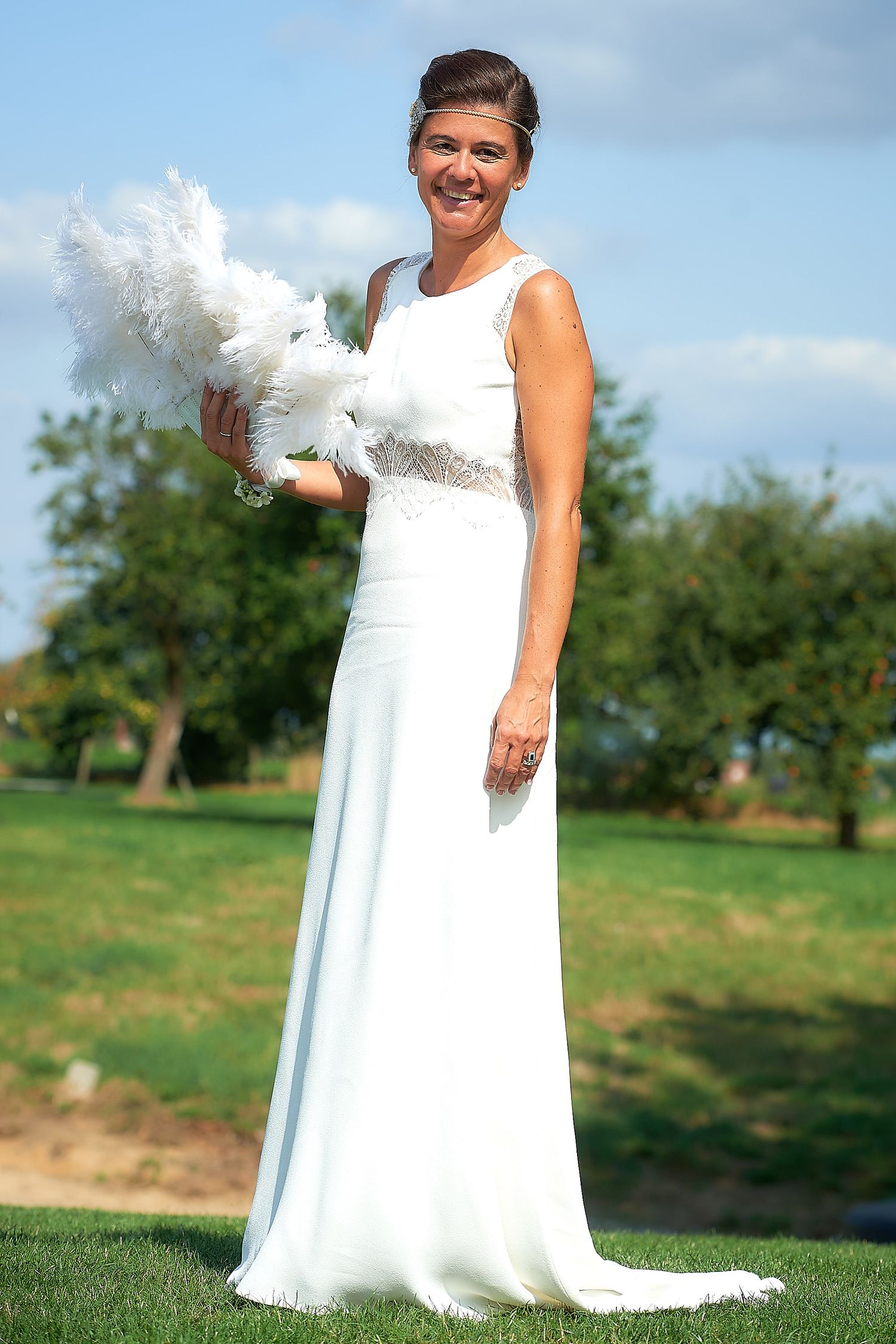 Smiling bride in elegant white gown holding white feathered bouquet outdoors on green lawn with trees and blue sky.