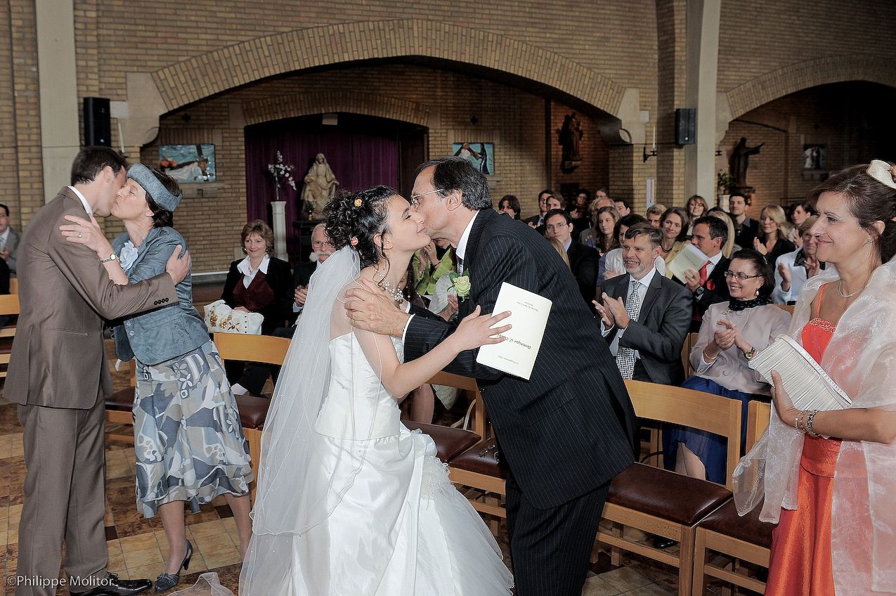 Bride and groom share a kiss at the altar during wedding ceremony as guests applaud in decorated church.