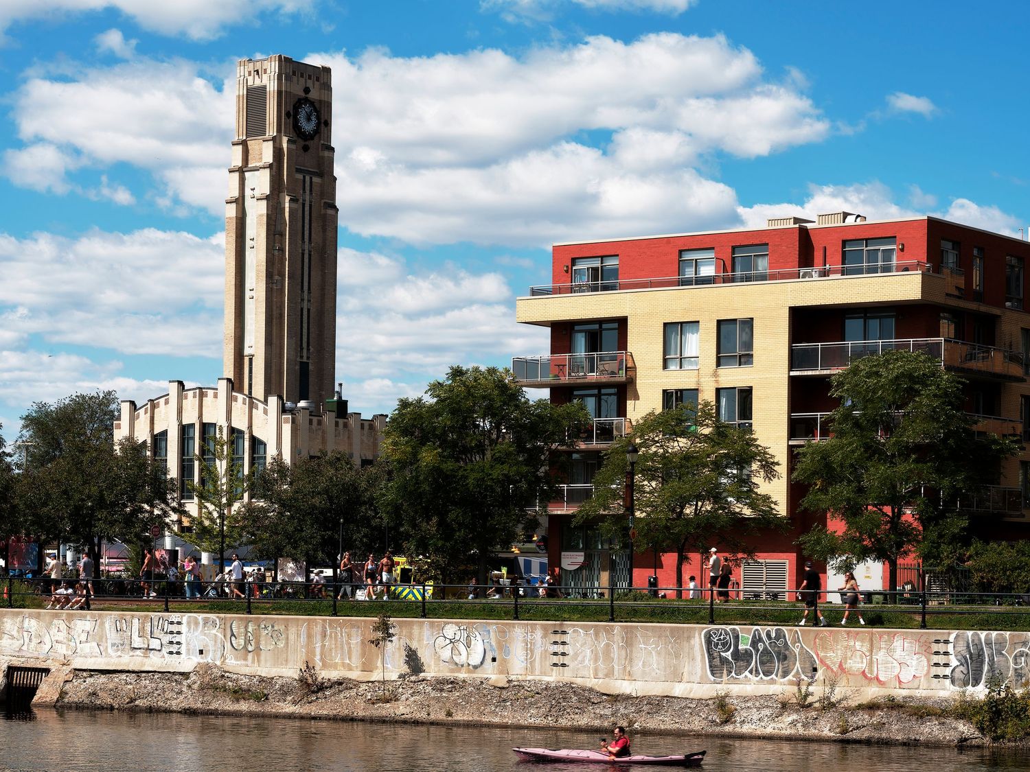 Art deco clock tower and modern red building along a urban canal with kayakers on a sunny day.