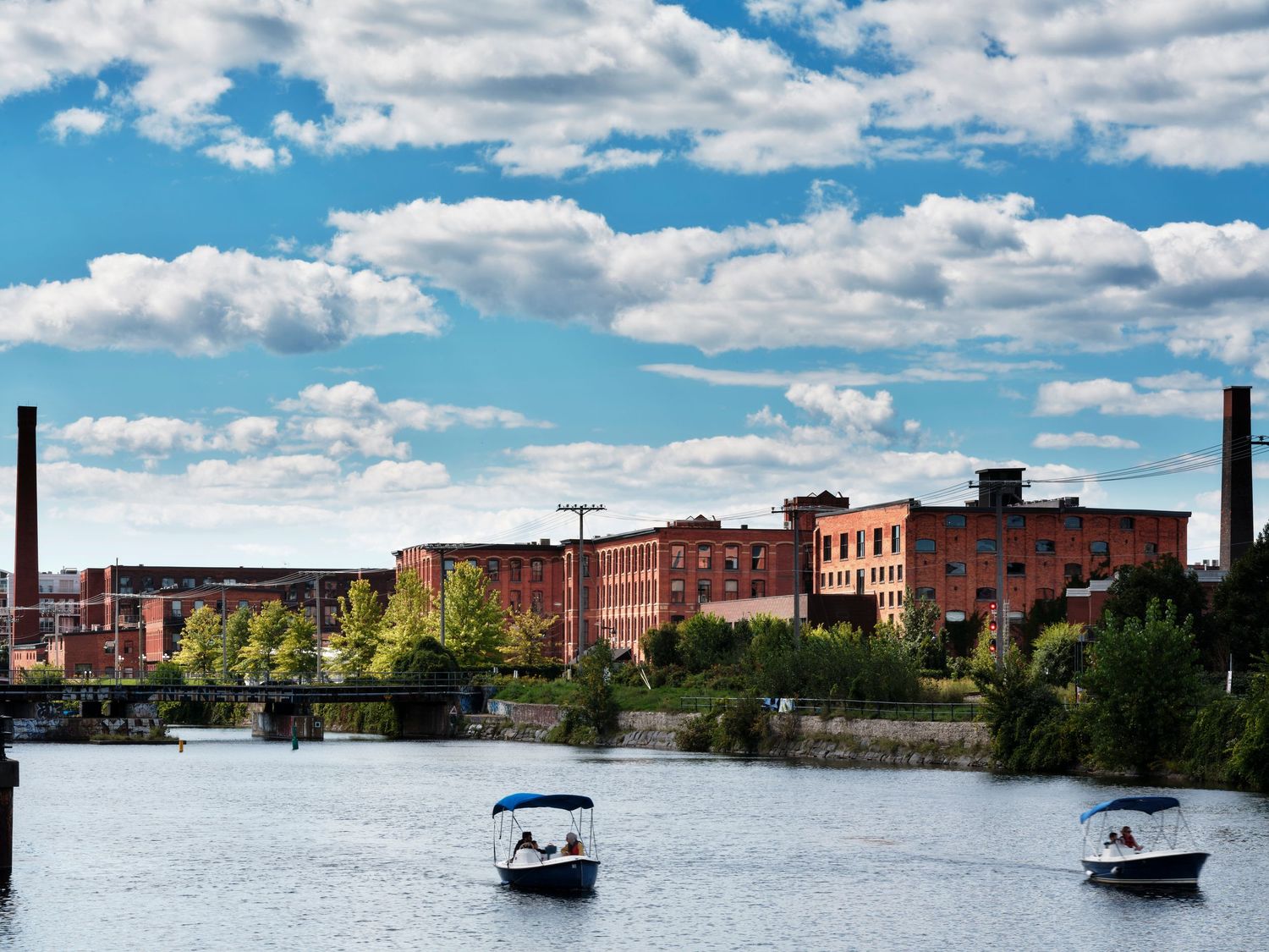 Two boats cruise along a river beside historic red brick mill buildings under a blue sky with fluffy white clouds.