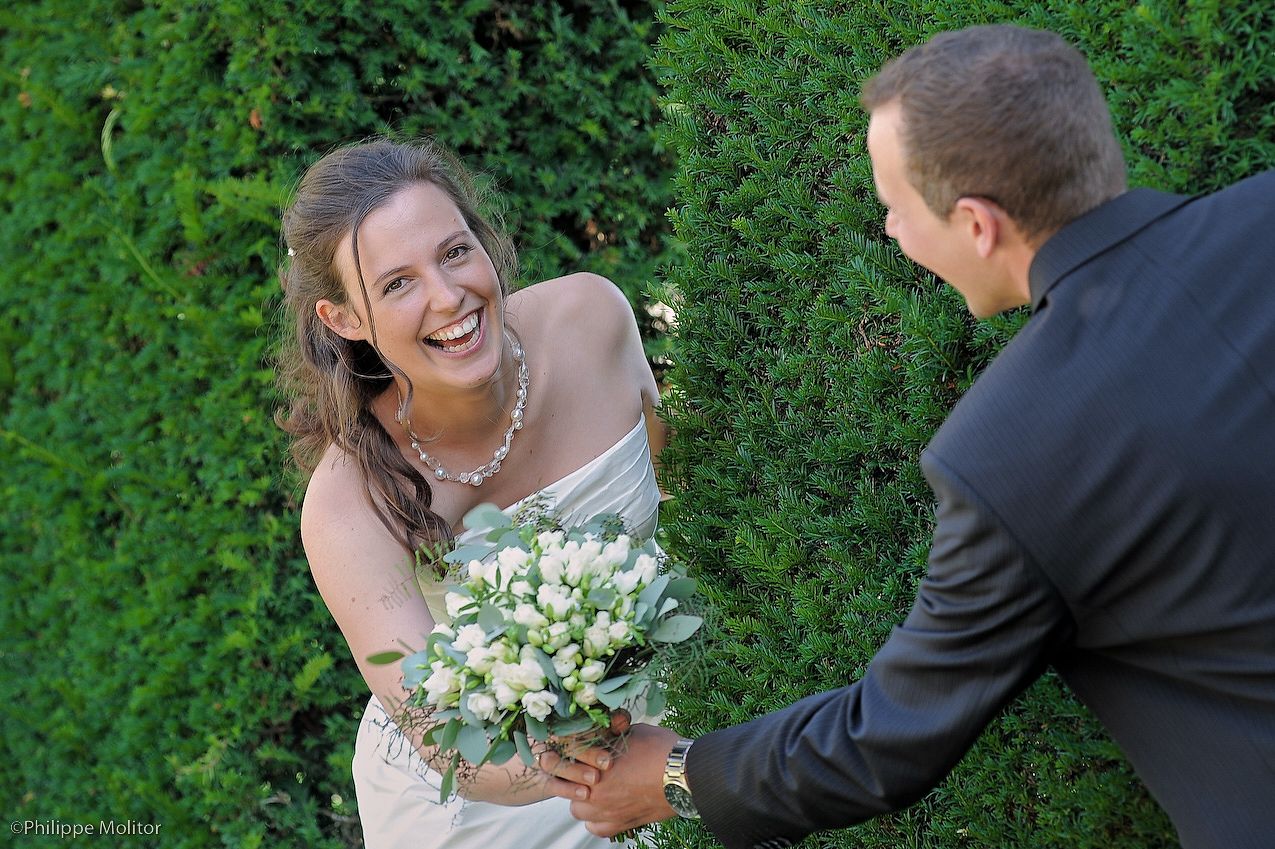 Bride laughing joyfully while holding white bouquet with groom in black suit, standing before lush green hedge backdrop.