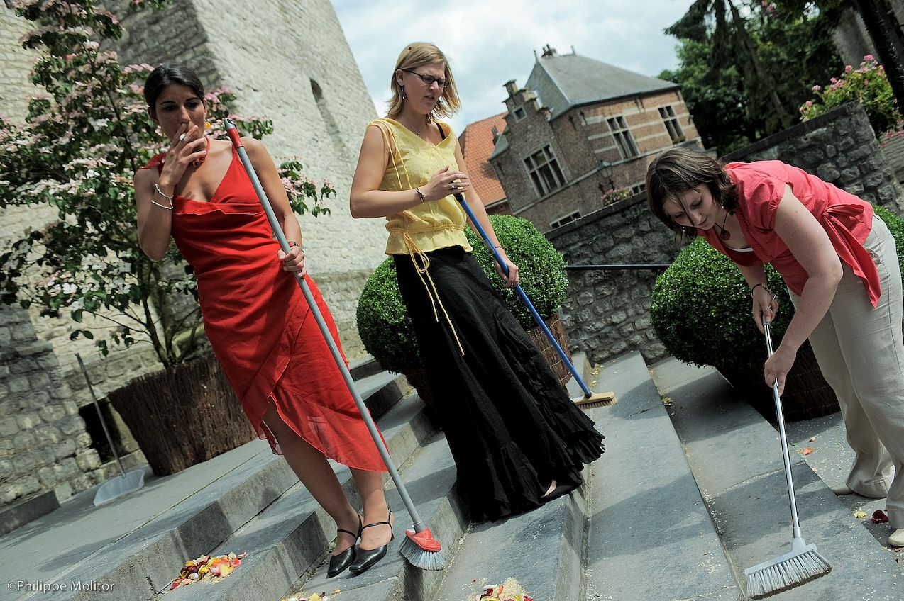 Three women in elegant dresses playfully sweeping outdoor stone steps near a historic building.