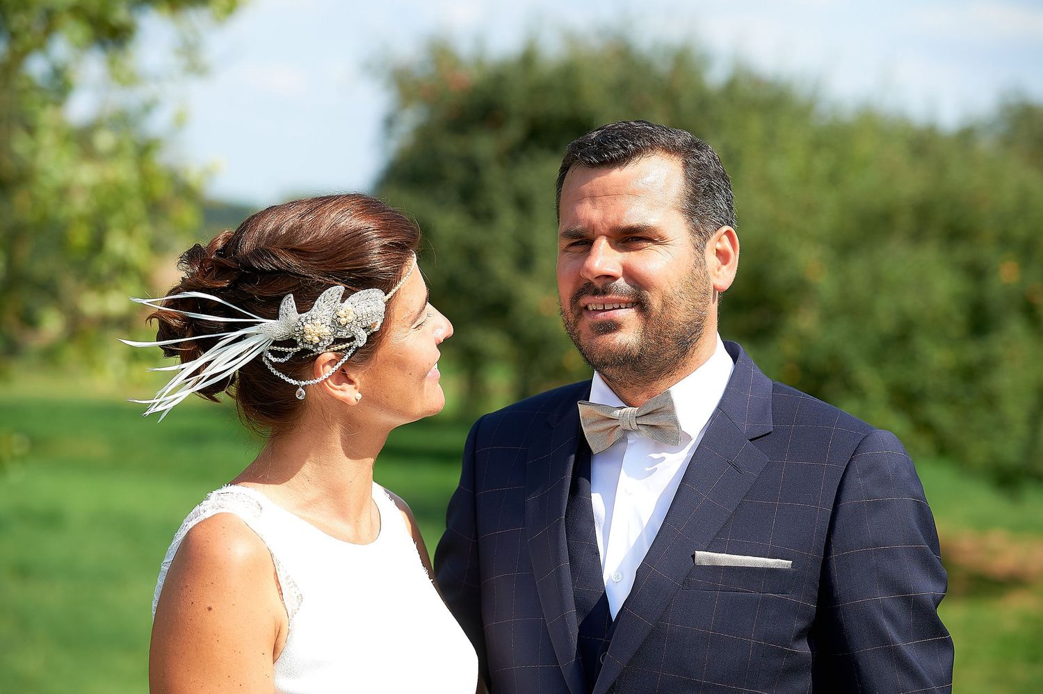 Bride and groom posing outdoors on their wedding day, woman in white dress with feather hair accessory.