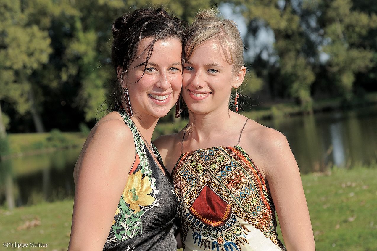Two smiling young women in colorful summer dresses posing together outdoors near a green park with trees and a pond.