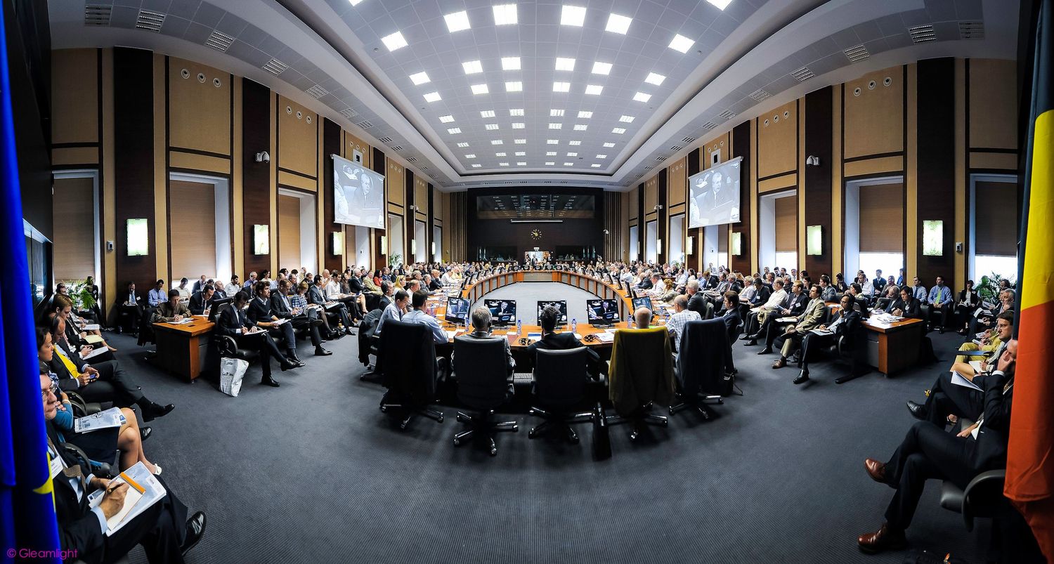Wide-angle view of dark conference room with illuminated ceiling grid and seated attendees.