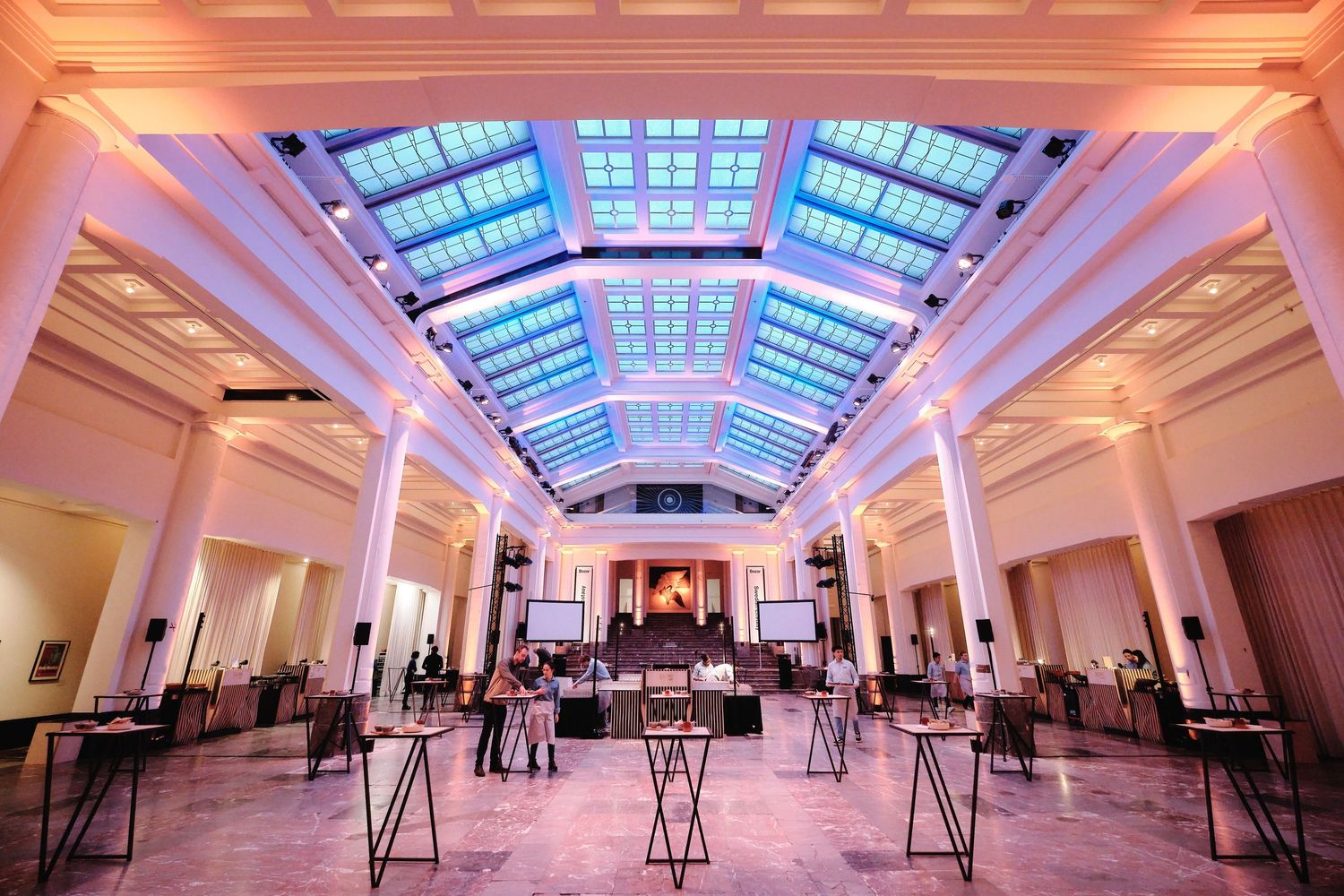 Modern convention center hall with skylight panels and white columns illuminated in pink lighting.