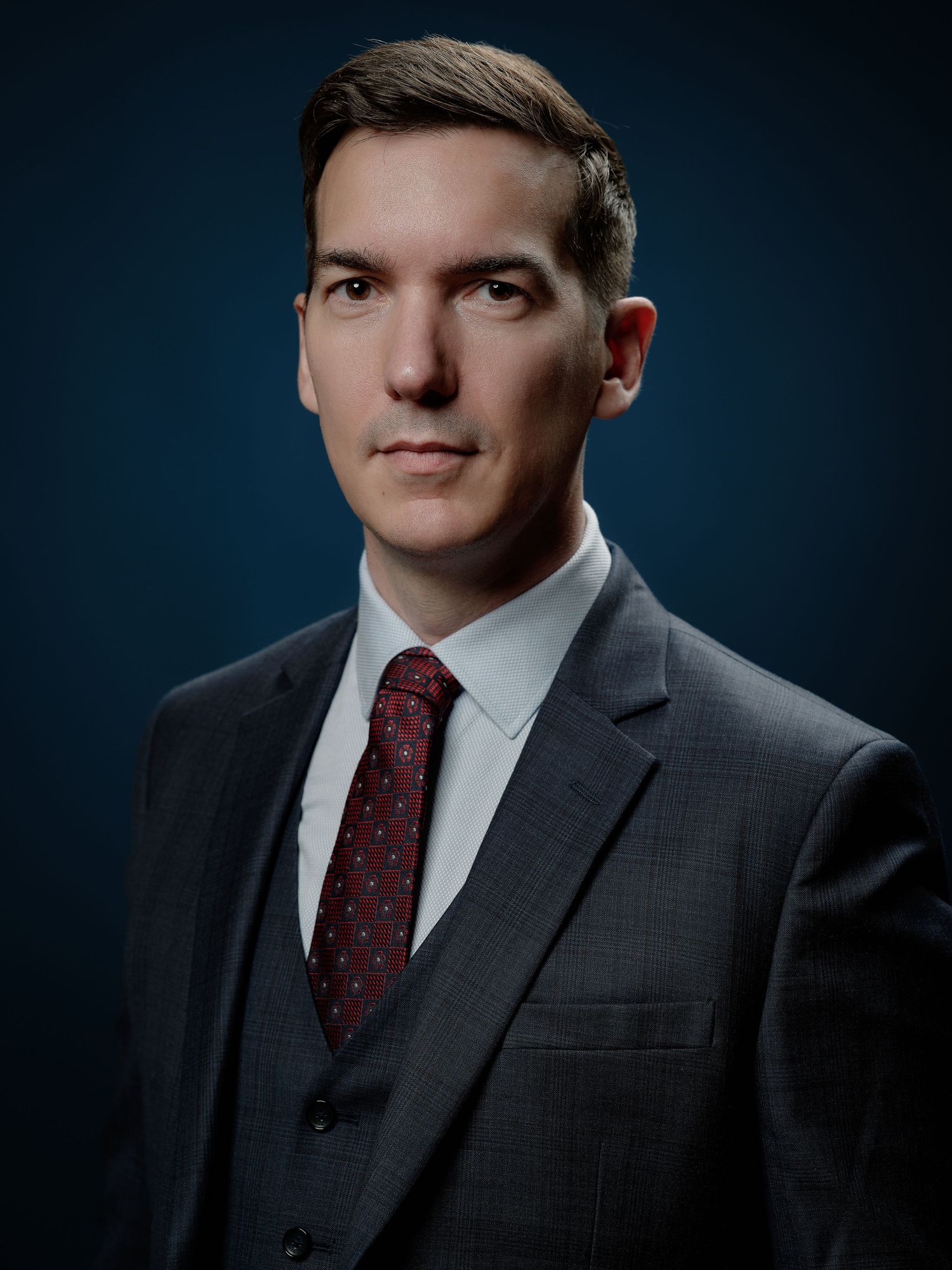 Un homme professionnel en costume gris et cravate rouge pose pour un portrait d'affaires formel sur fond bleu foncé. Portrait entreprise.