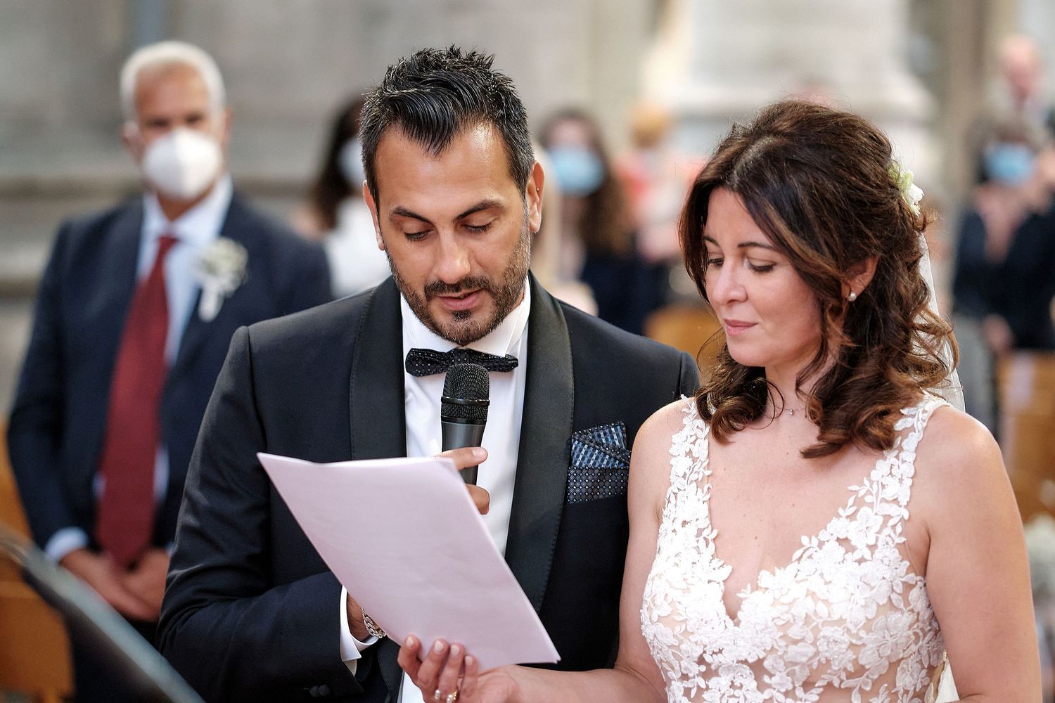 Bride and groom reading wedding vows from paper during ceremony, guests wearing masks in background.