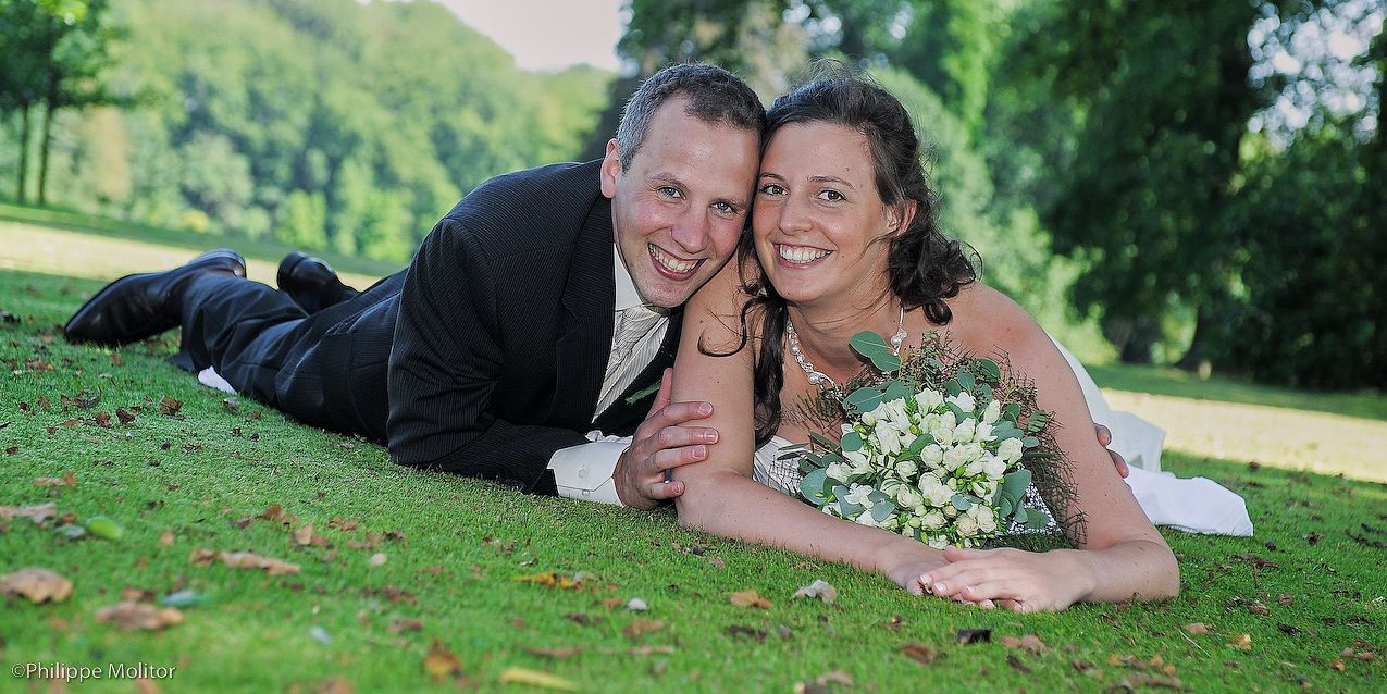 Smiling bride and groom lying on green grass in a park, with bride holding a white wedding bouquet.