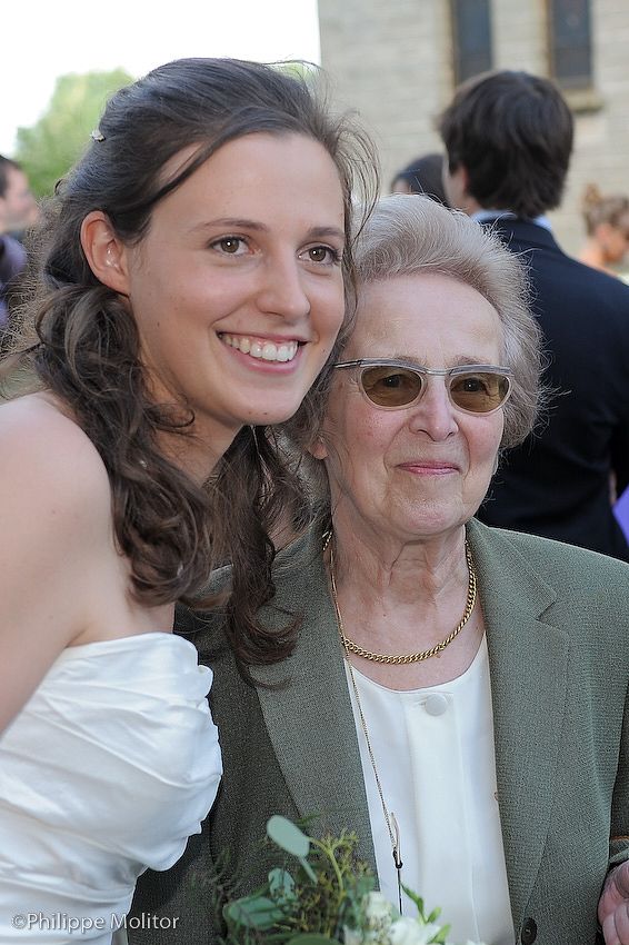 Young bride in white dress smiles with elderly woman in green jacket and sunglasses at outdoor wedding.