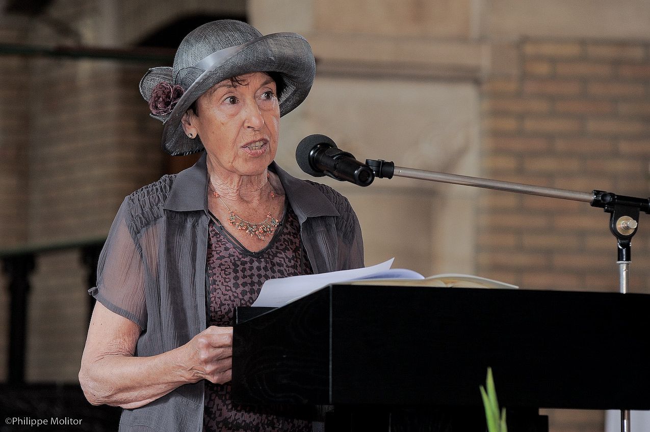 Elderly woman in grey hat speaking at podium with microphone during formal ceremony.