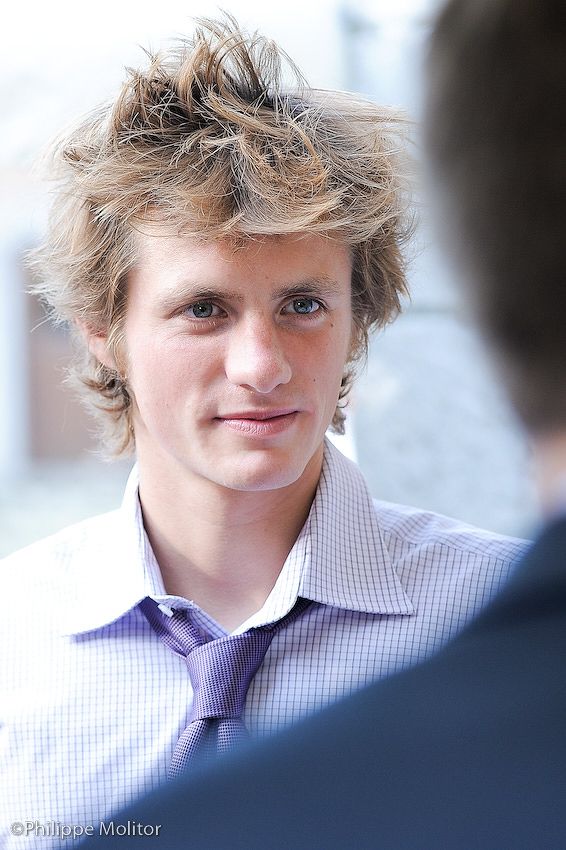 Young man with tousled blonde hair wearing a purple shirt and tie at a formal event.