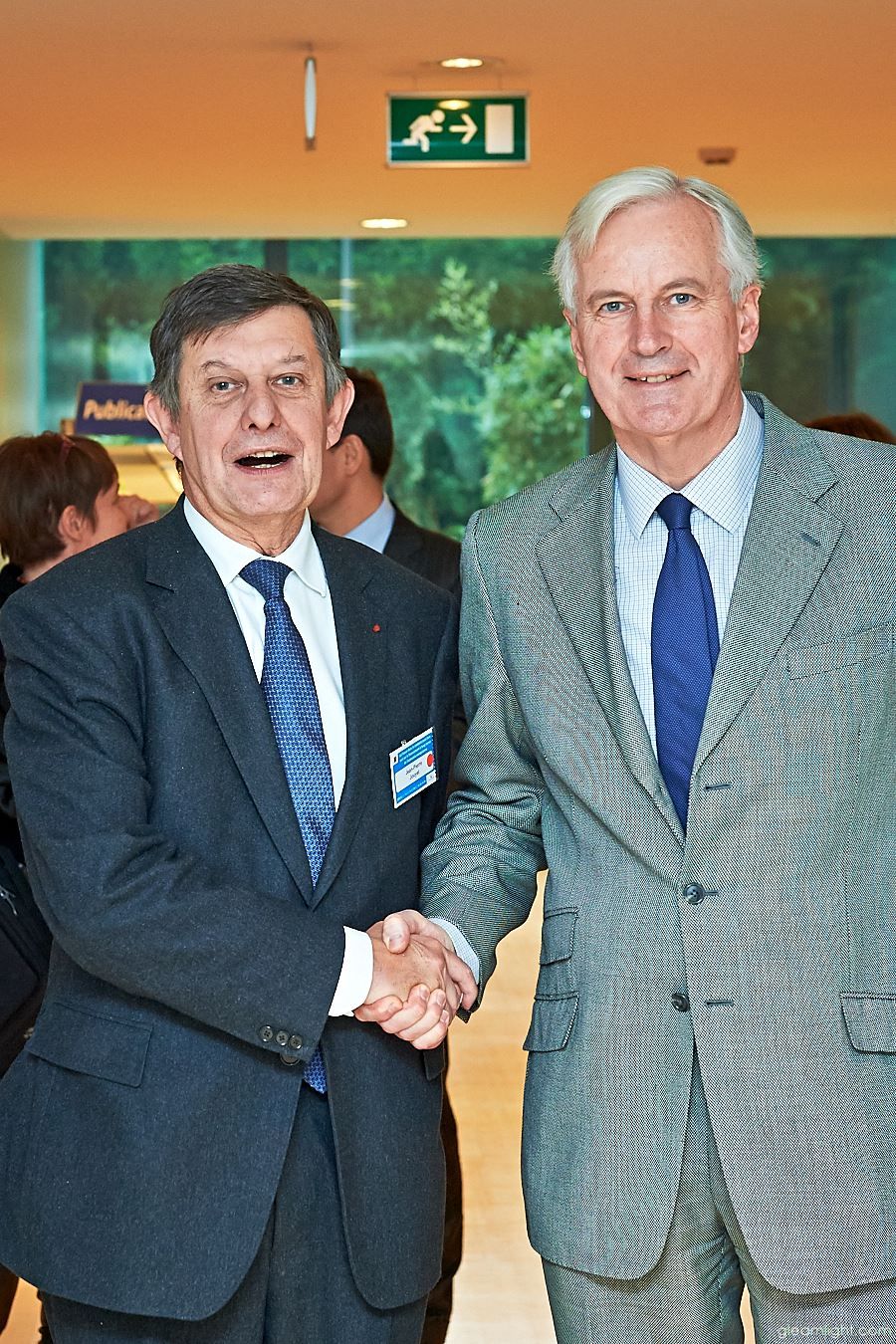 Two executives in business suits shaking hands in an office hallway.