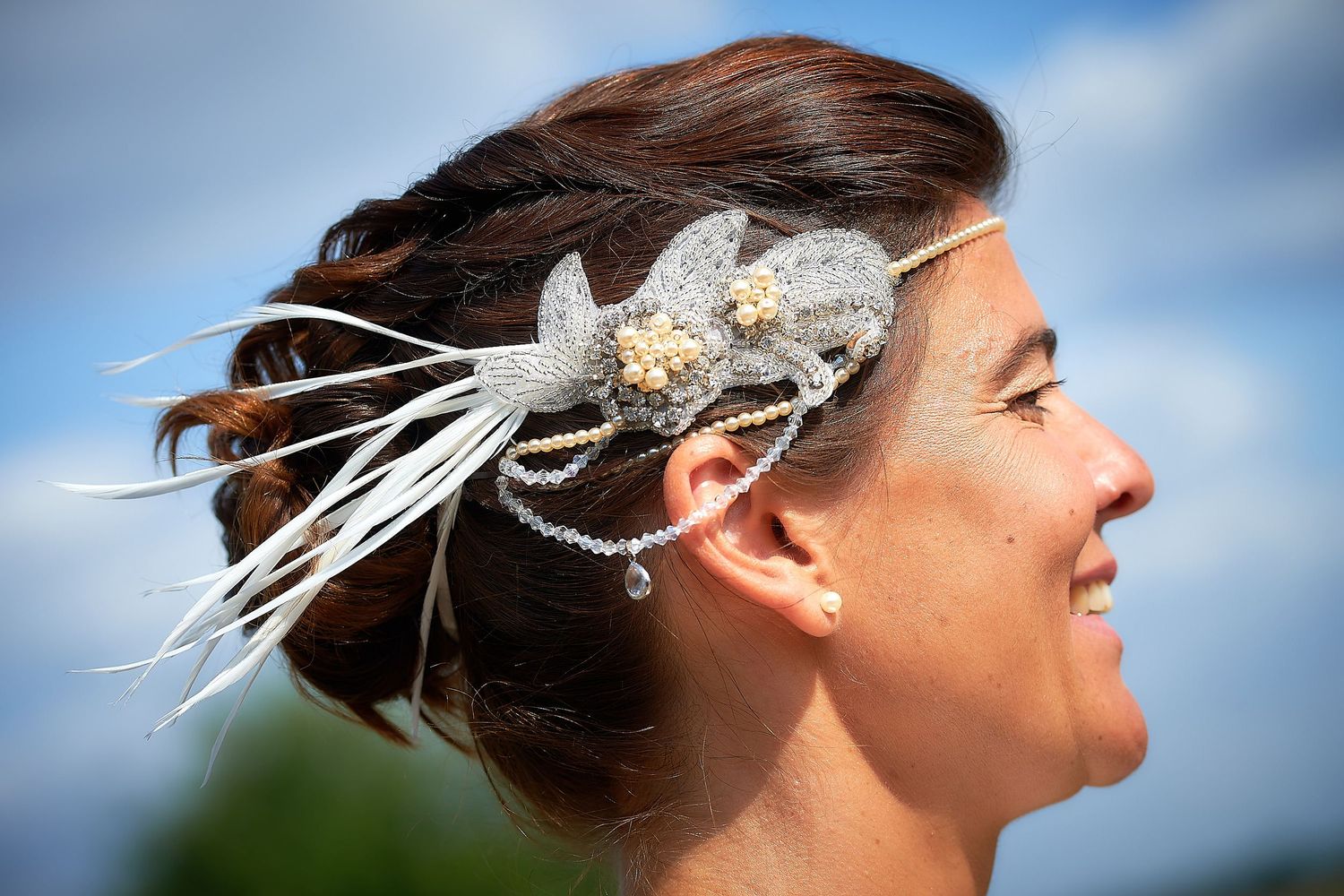 Smiling bride wearing elegant feather and pearl hair accessory with crystal chain against blue sky.