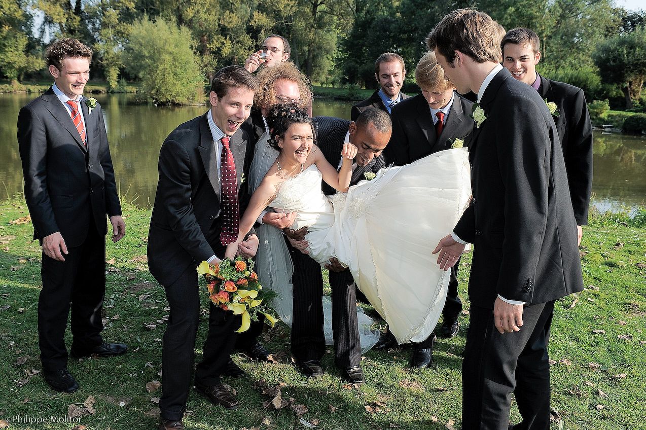 Groomsmen lifting the bride in a white wedding dress outdoors near a pond during a fun wedding party photo.