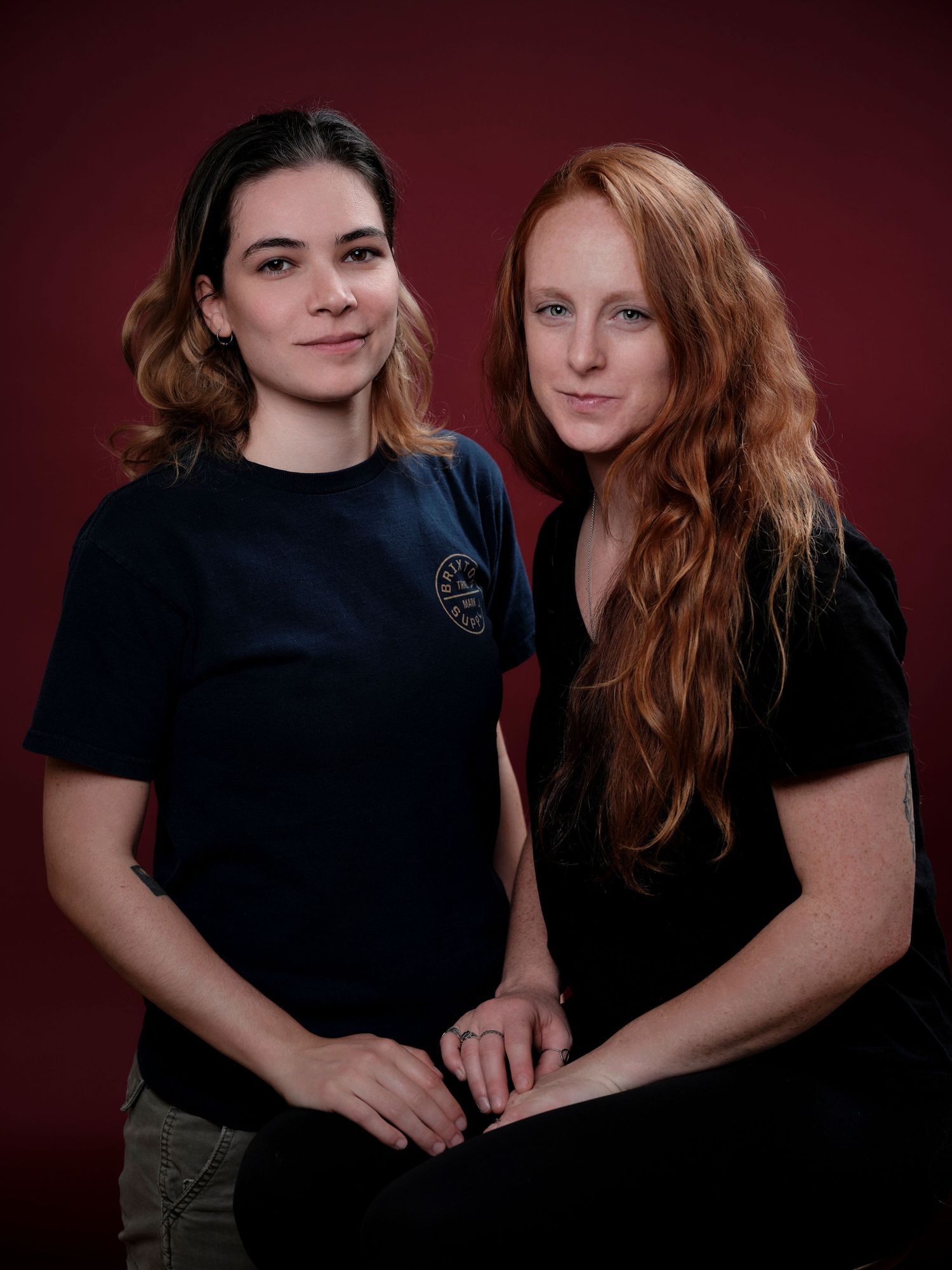 Two women posing together against a dark red background, one with dark hair and one with long red hair, both wearing black t-shirts.