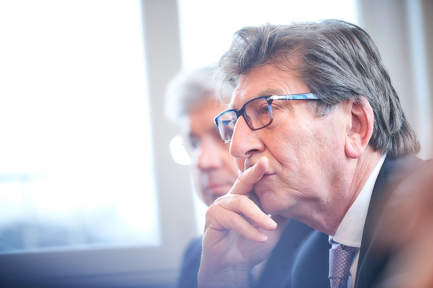 A thoughtful person in glasses and business attire listens intently during a meeting.