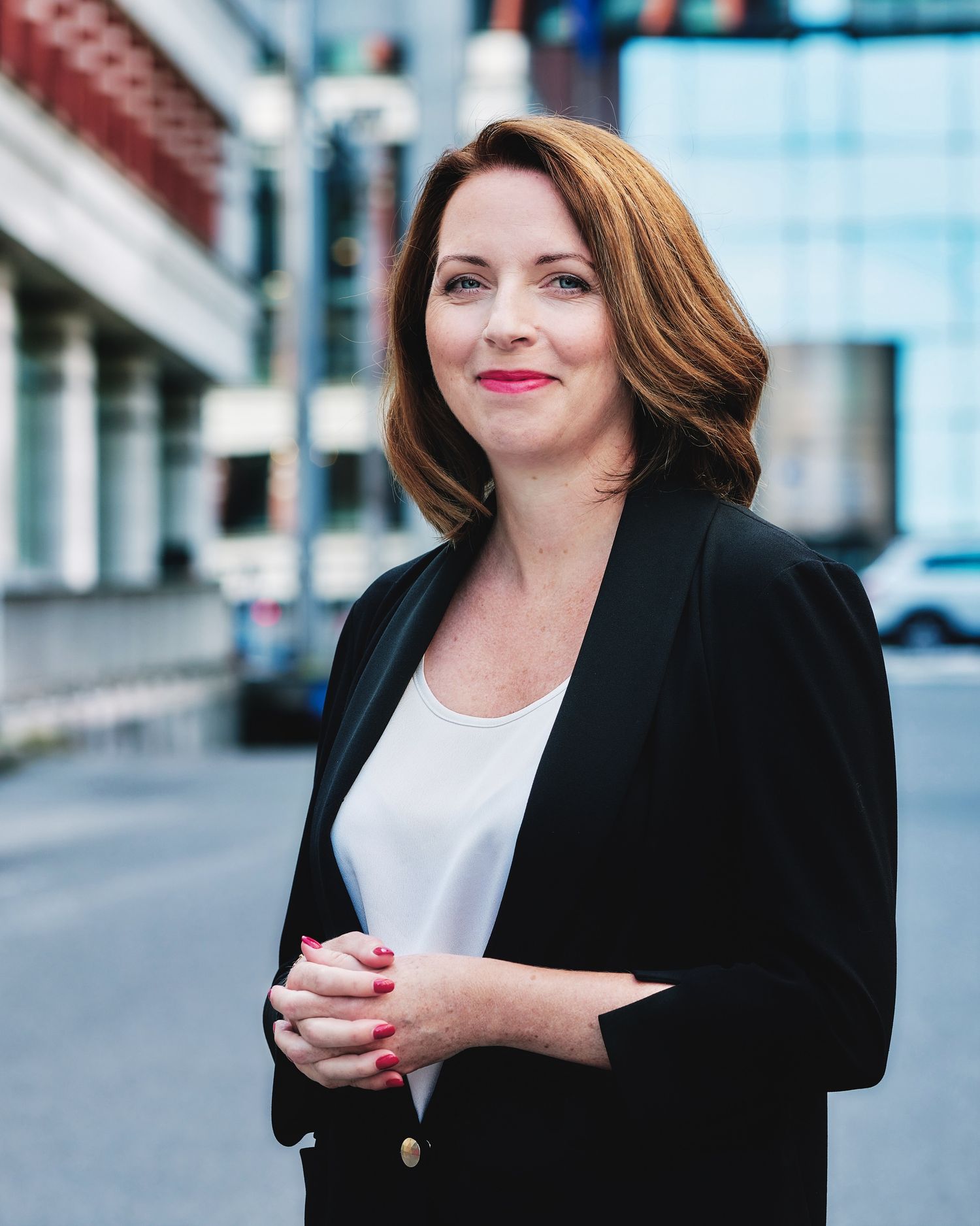 Professional portrait of person in black blazer and white top against urban background.