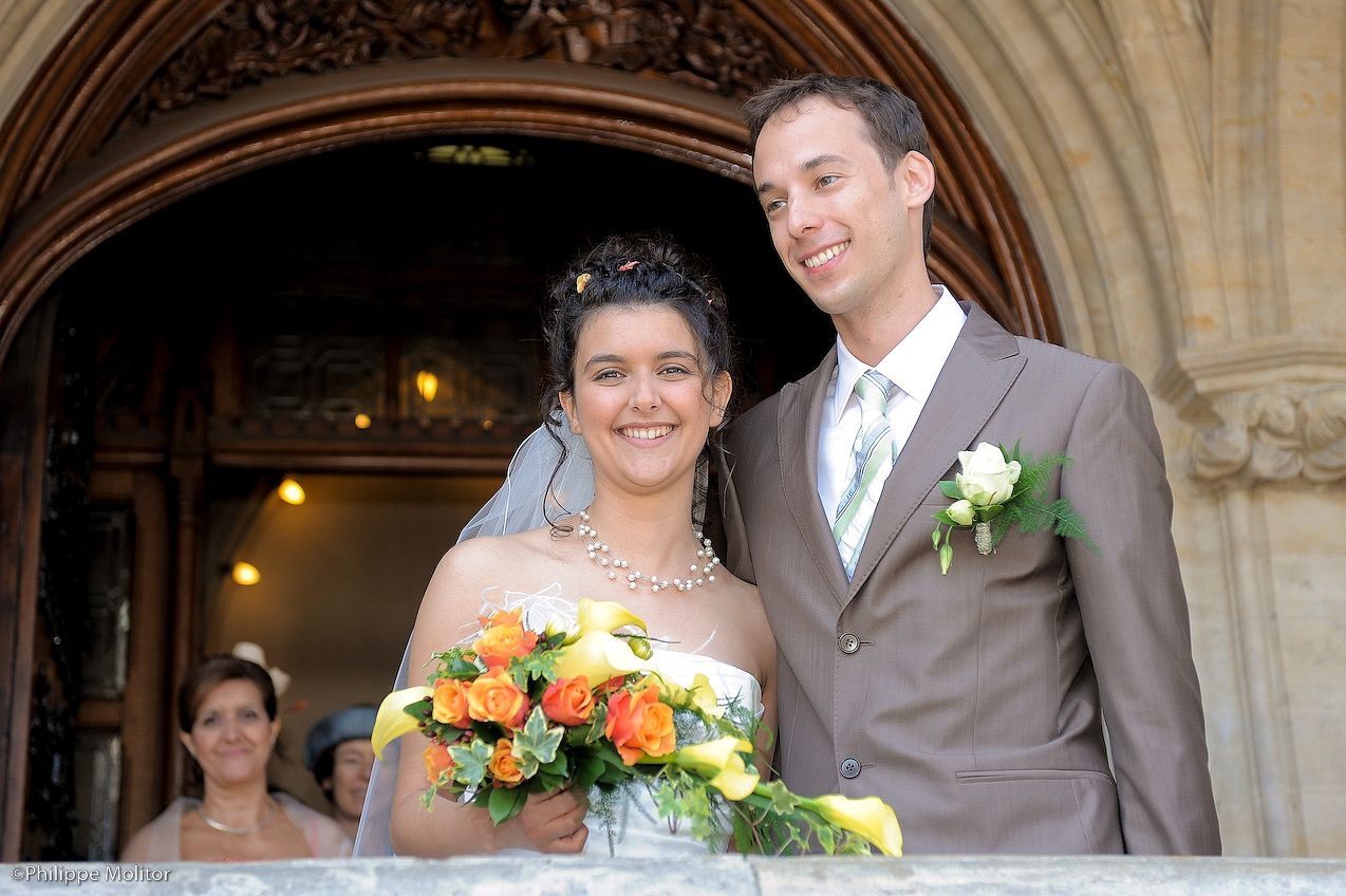 Happy bride and groom posing at church entrance, bride holding colorful bouquet in wedding attire.