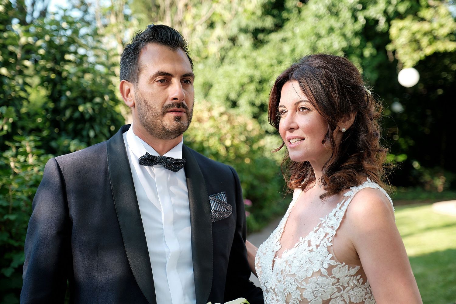 Elegant couple in wedding attire posing outdoors, groom in tuxedo and bride in lace dress, surrounded by greenery.