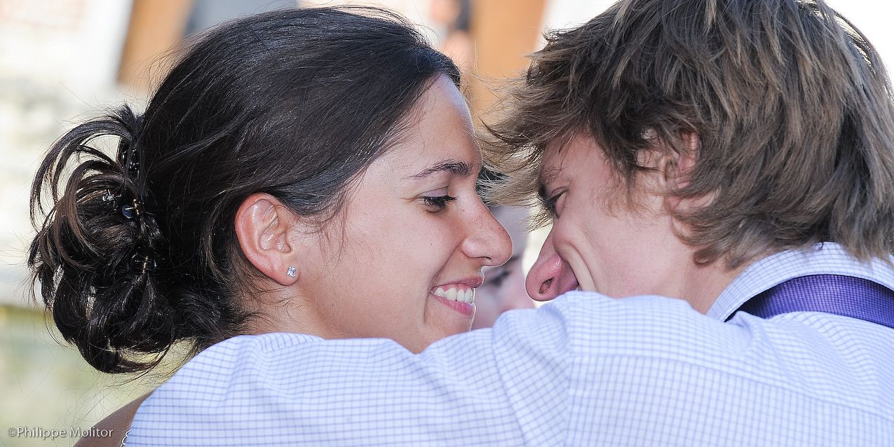 A smiling couple shares a tender romantic moment, leaning close together outdoors at what appears to be a wedding.