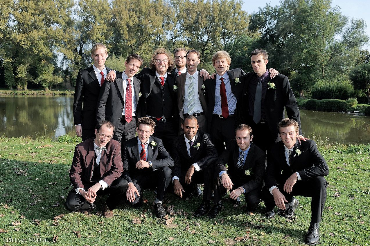 Group of groomsmen in suits pose together outdoors near a pond for a wedding photo.