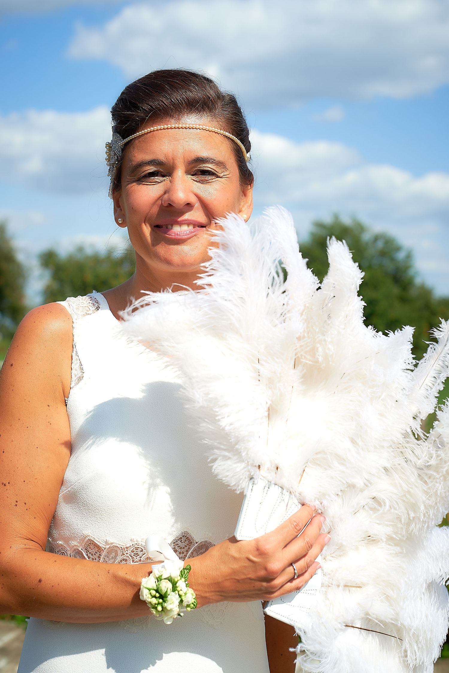 Smiling bride in white dress holding large white feather bouquet outdoors on sunny day with blue sky background.