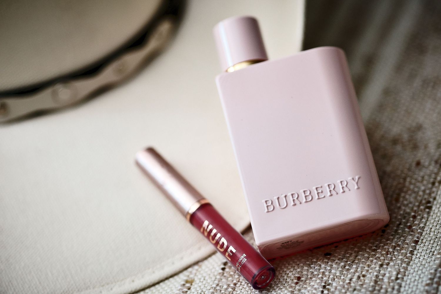 Burberry Her Elixir perfume bottle in blush pink beside a nude lip gloss on a neutral background.