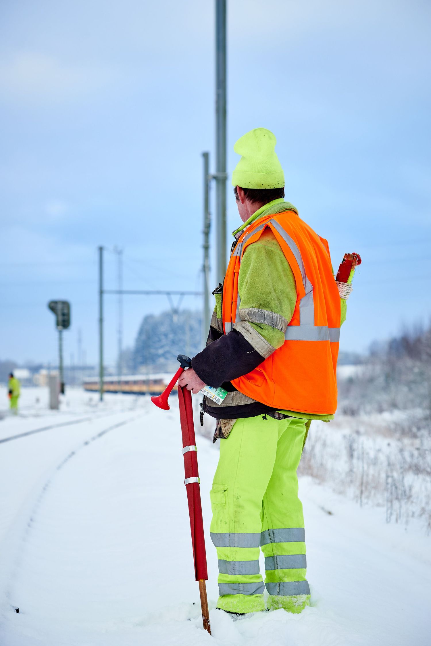 Railroad worker in bright safety gear stands with maintenance equipment on snowy tracks.
