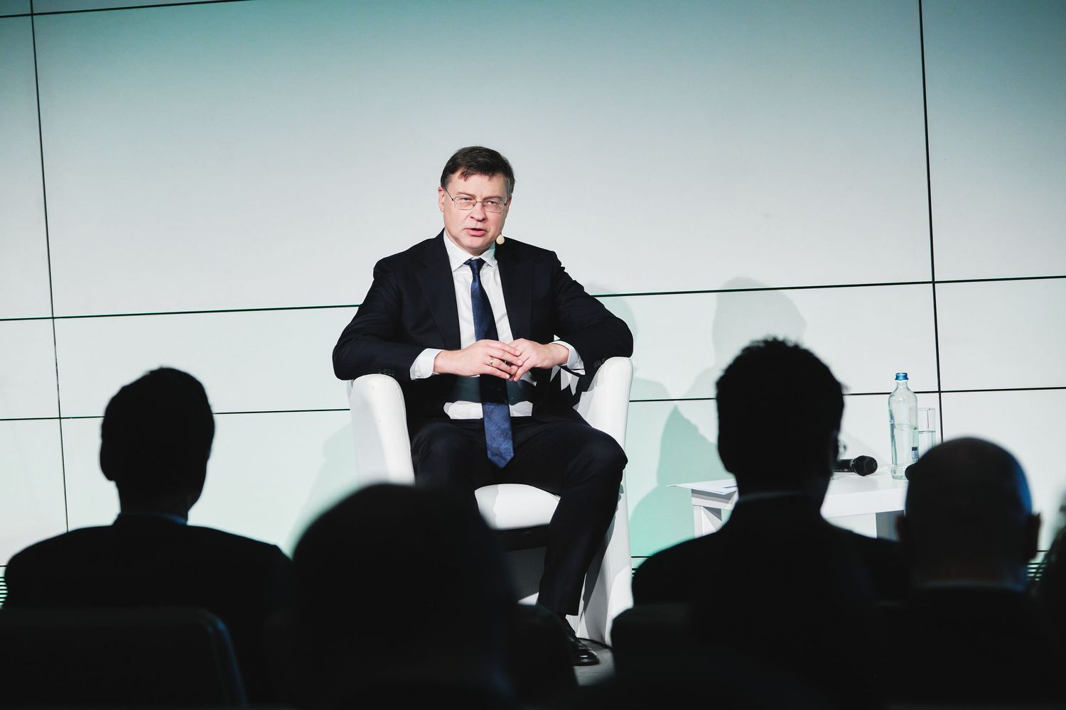 Speaker in dark suit addresses audience silhouetted in foreground during conference presentation.