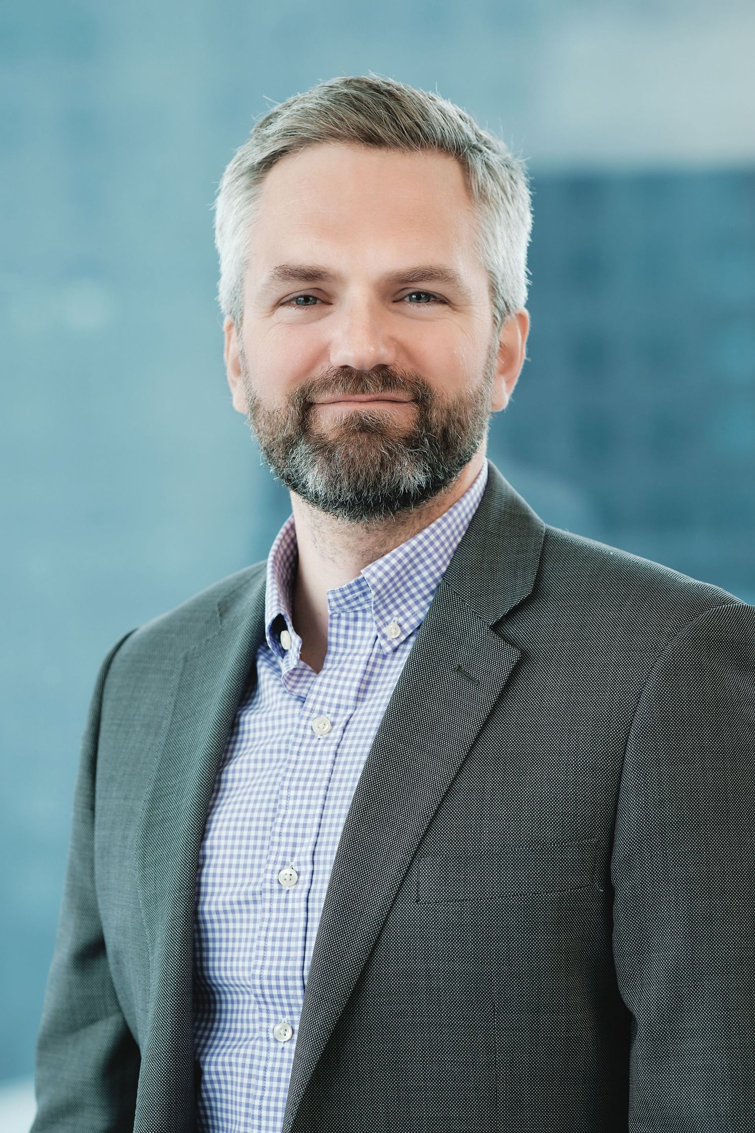 Professional portrait of an executive in grey suit jacket and checkered shirt with office background.