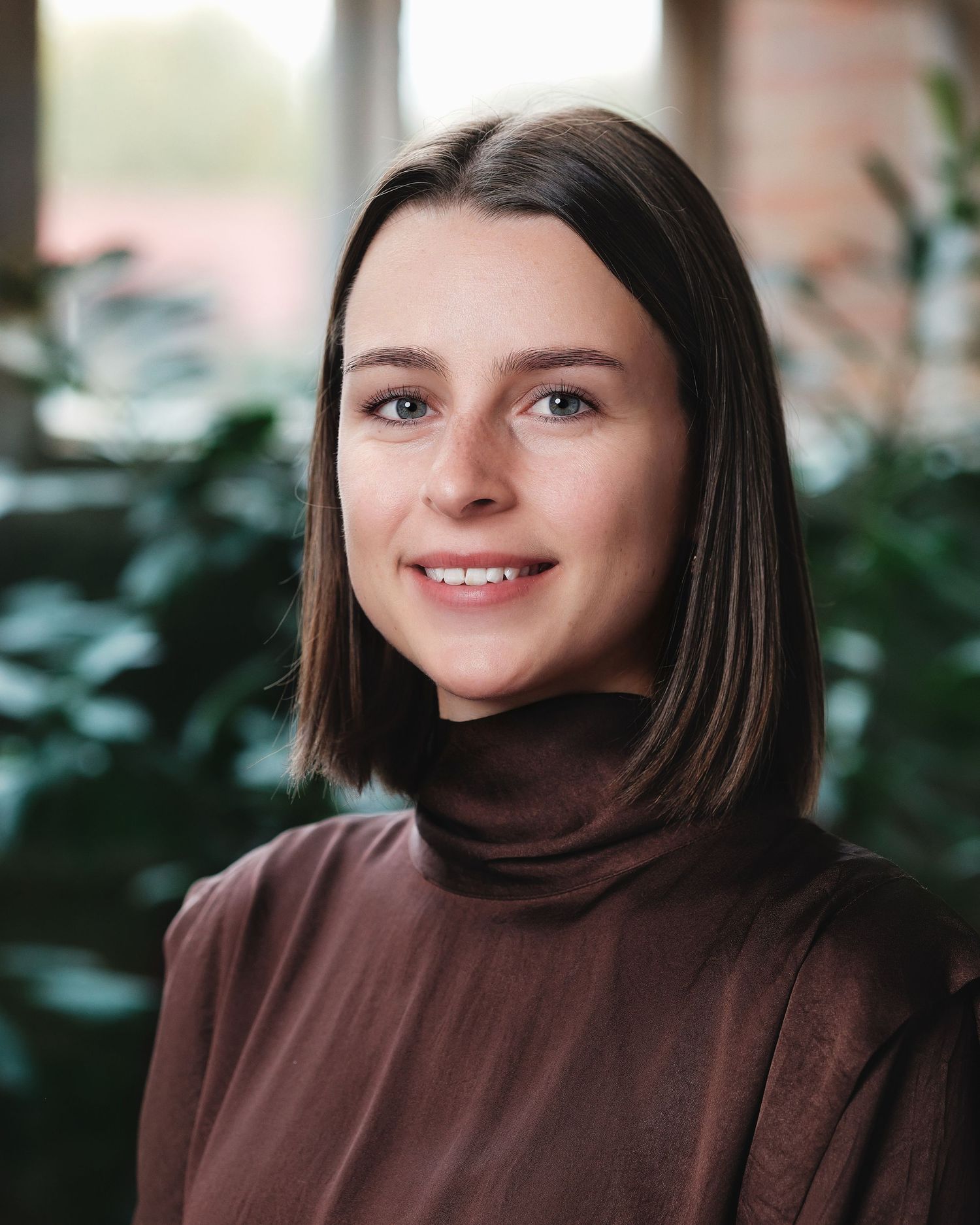 Professional wearing a brown turtleneck sweater in an office with plants in background.