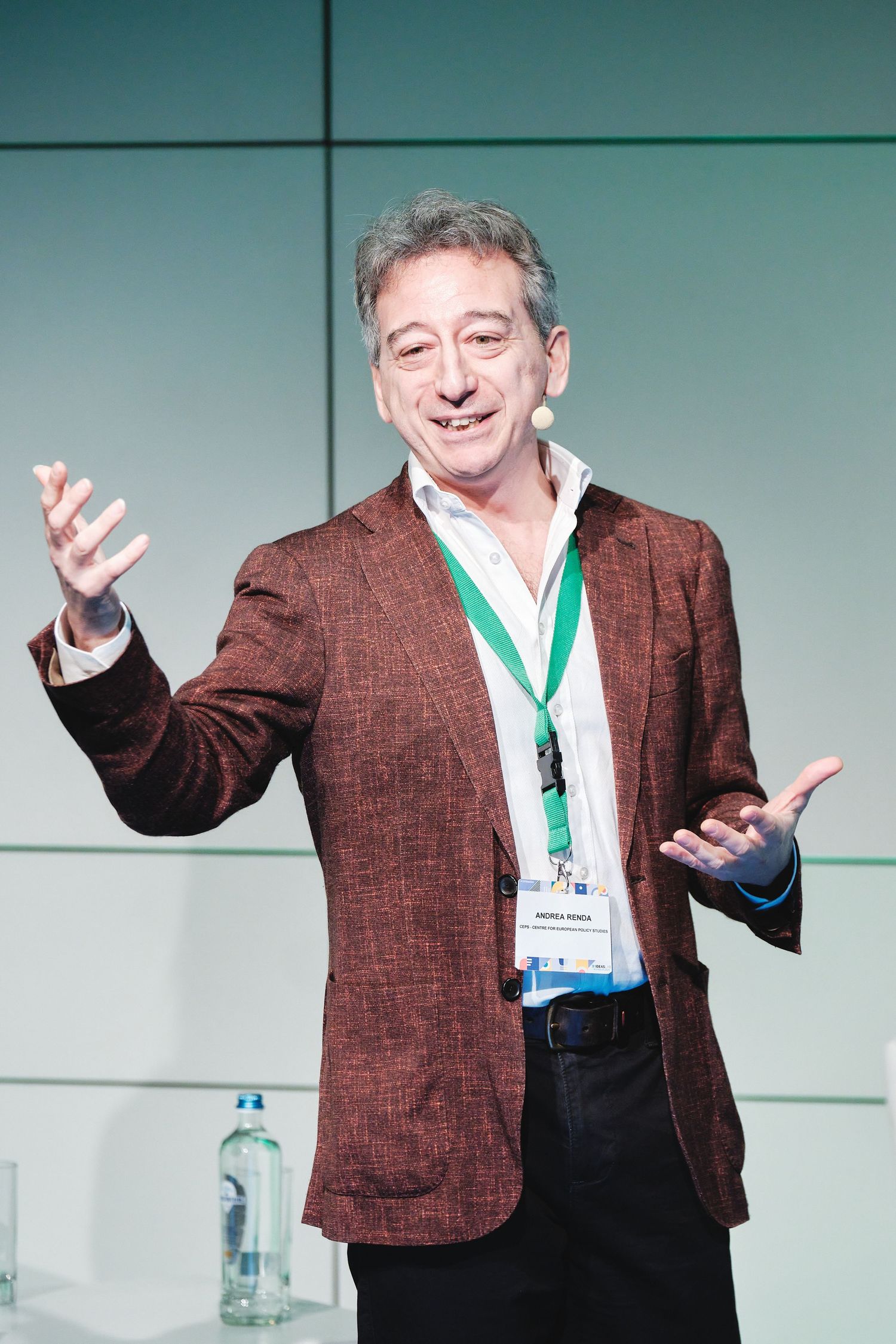 Speaker in brown blazer gestures enthusiastically while presenting at conference.