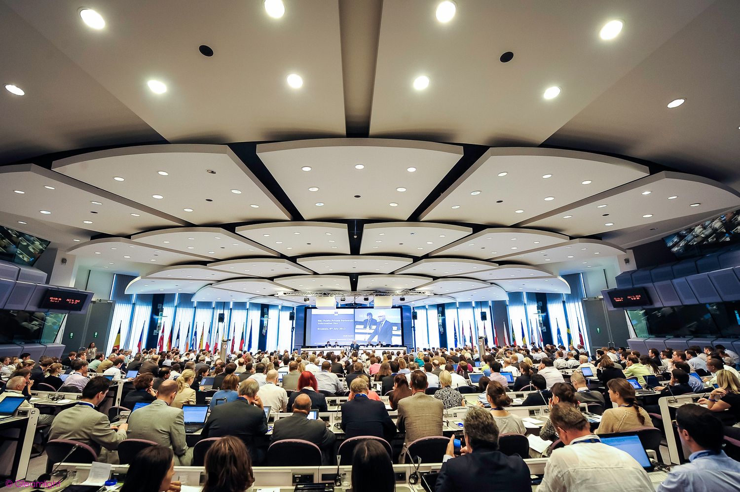 Panoramic view of a packed conference hall with rows of attendees under recessed lighting.