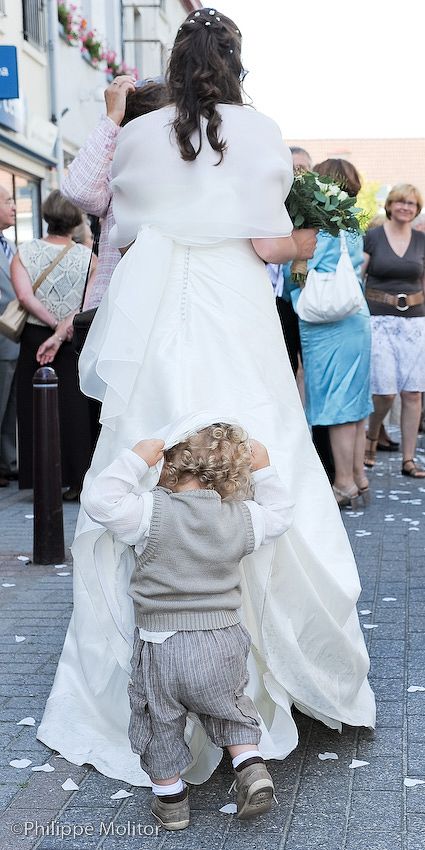 A toddler in a gray suit lifts a bride's white wedding gown train on a cobblestone street during an outdoor ceremony.