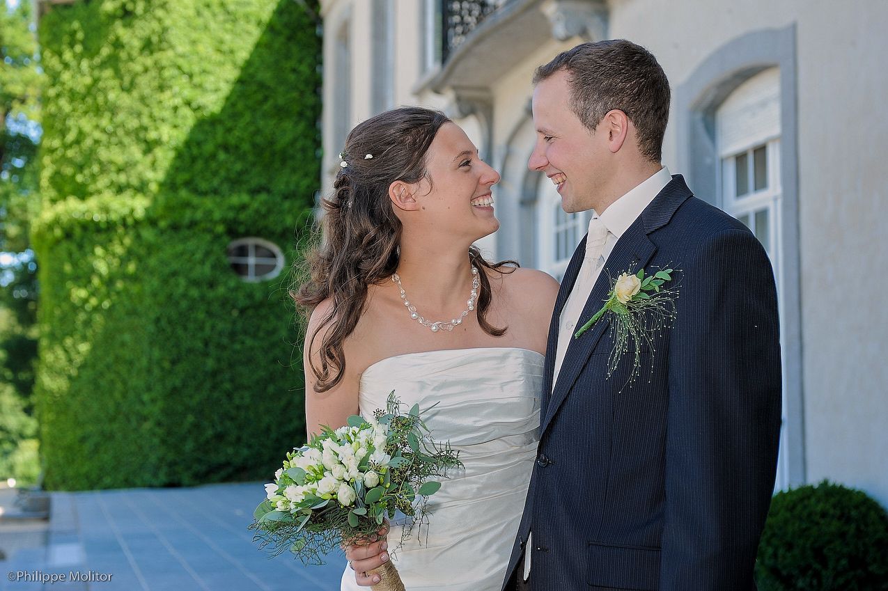 A smiling bride holding a white bouquet and groom in navy suit with boutonniere pose together outdoors at a wedding venue.